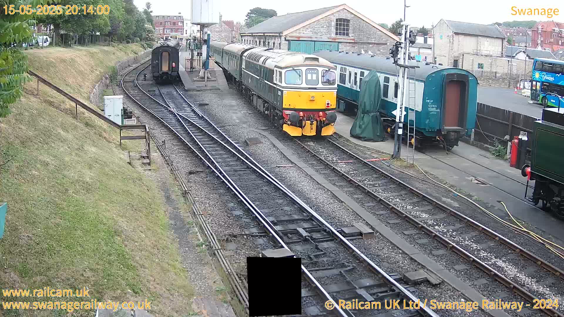 A train sits at a railway station on an overcast day, with other train cars and railway buildings visible.