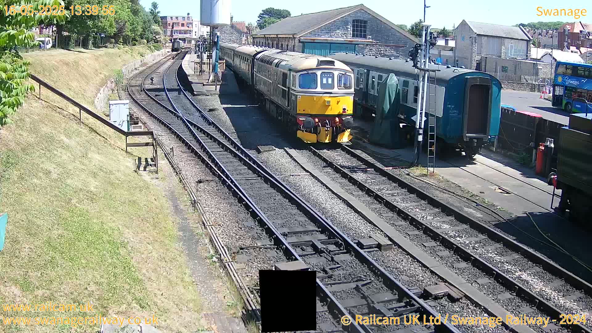 A yellow and dark-green diesel train is moving on railway tracks in bright sunlight, alongside other railway cars and a building.
