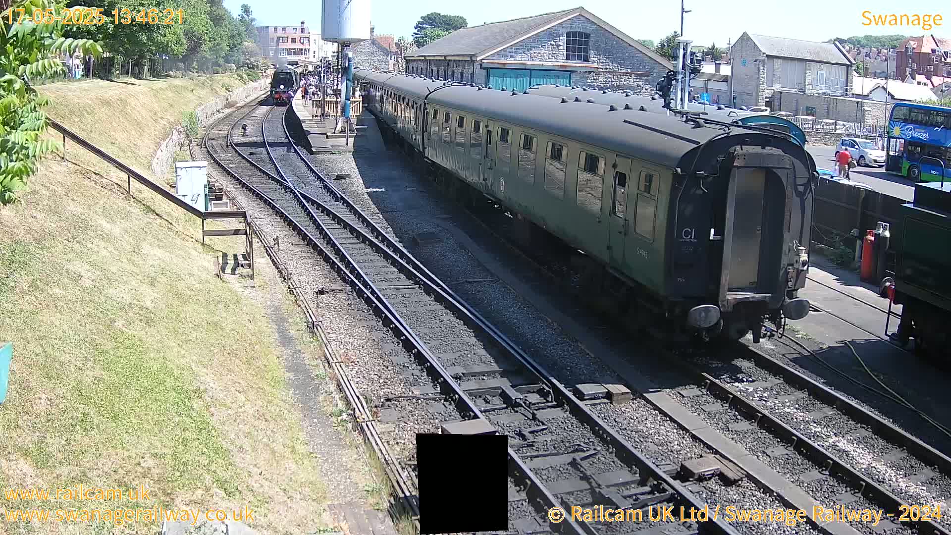 A long green passenger train is at a station on a sunny day, with another train visible further down the tracks.
