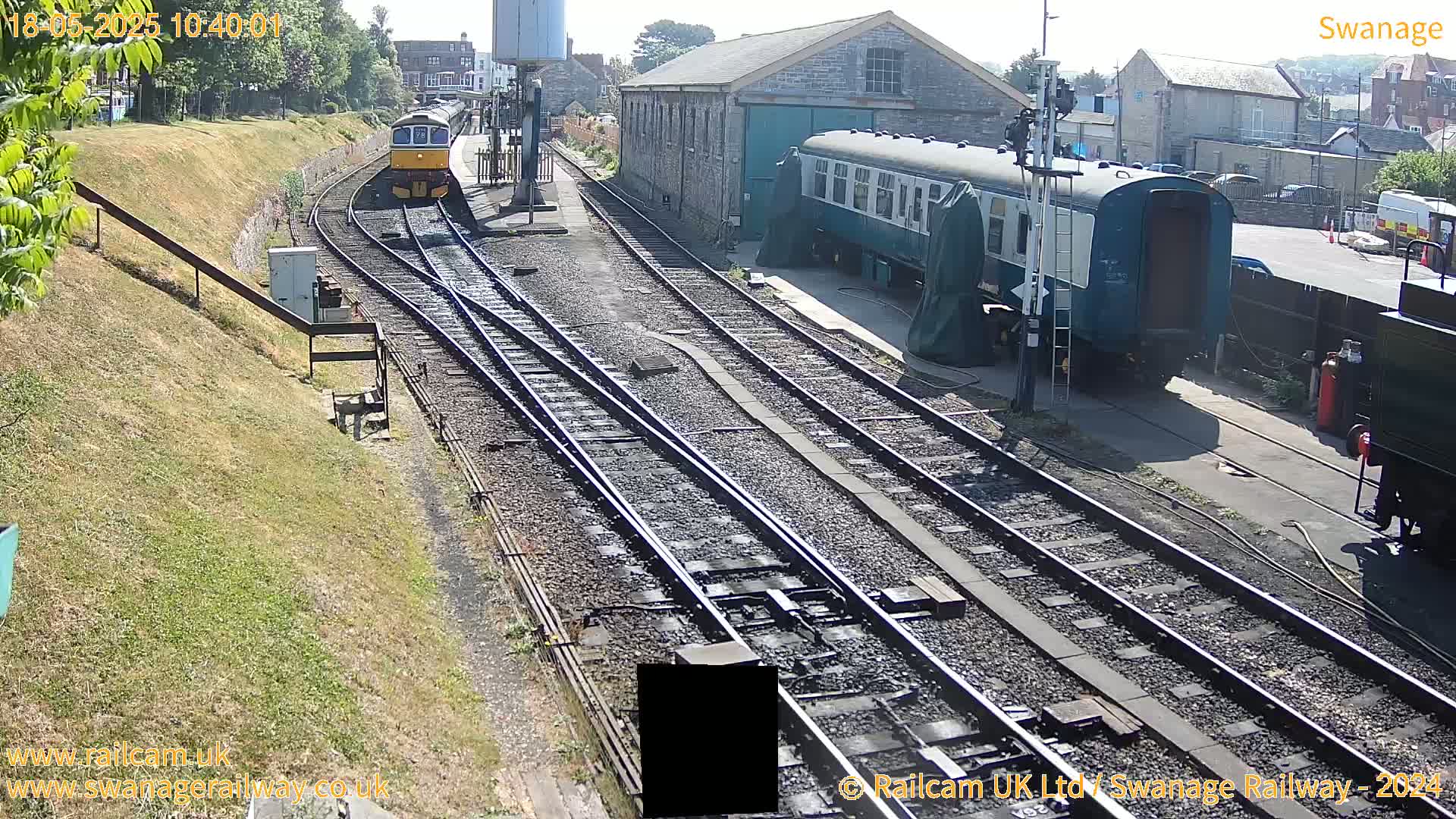 A sunny day at a railway station shows multiple train cars, some on tracks and others parked near a stone building.