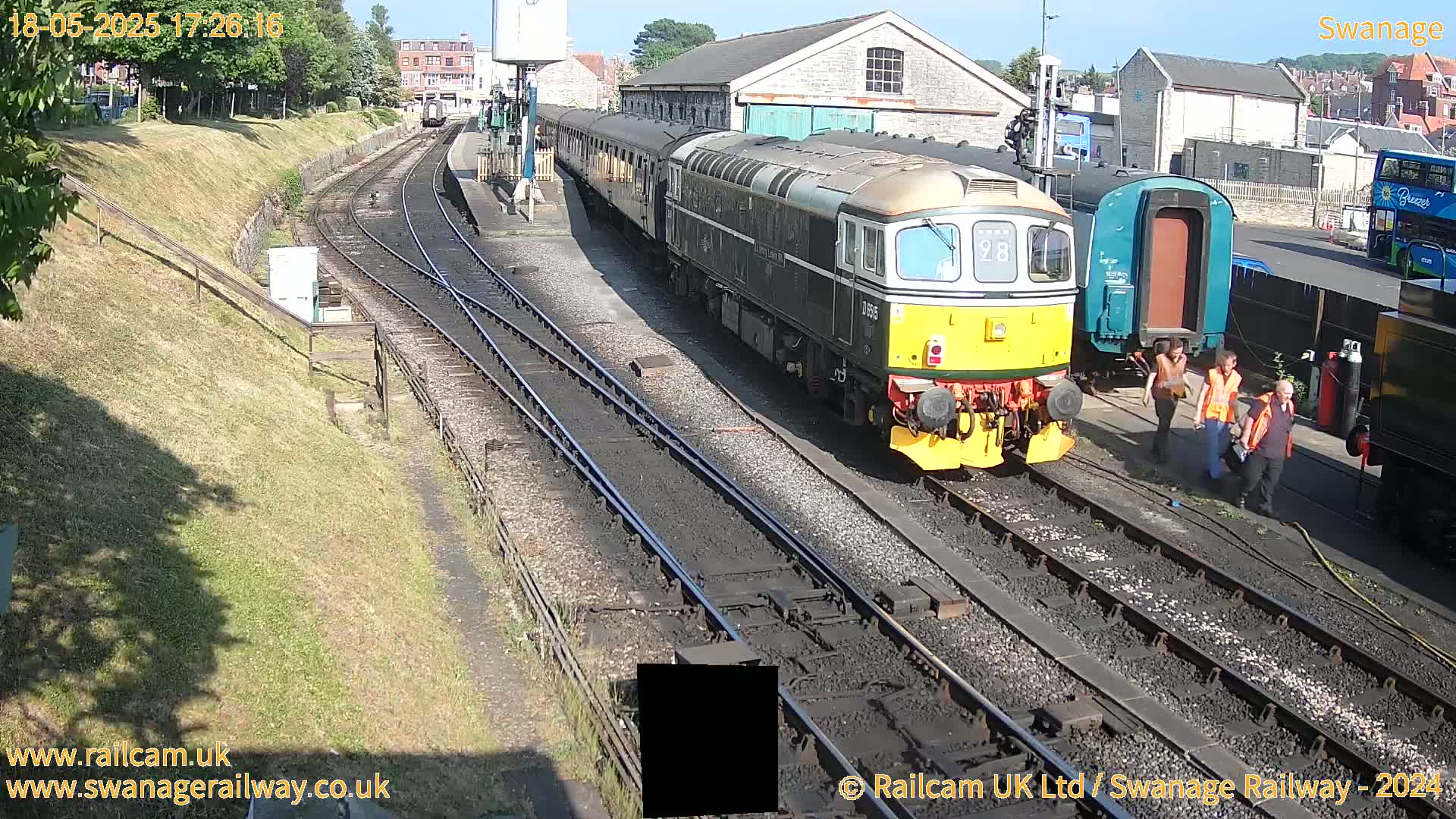 A diesel train is stopped at a railway station on a sunny day, with several workers standing nearby.