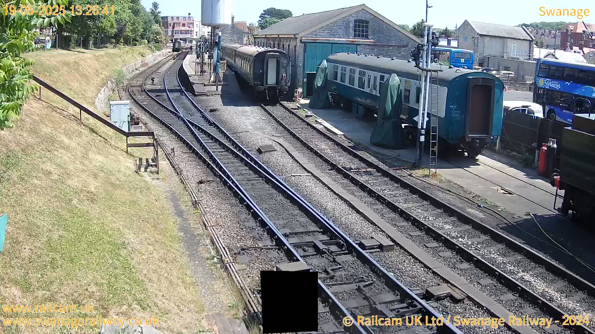 Several railway carriages are parked along train tracks on a sunny day near a railway station building.