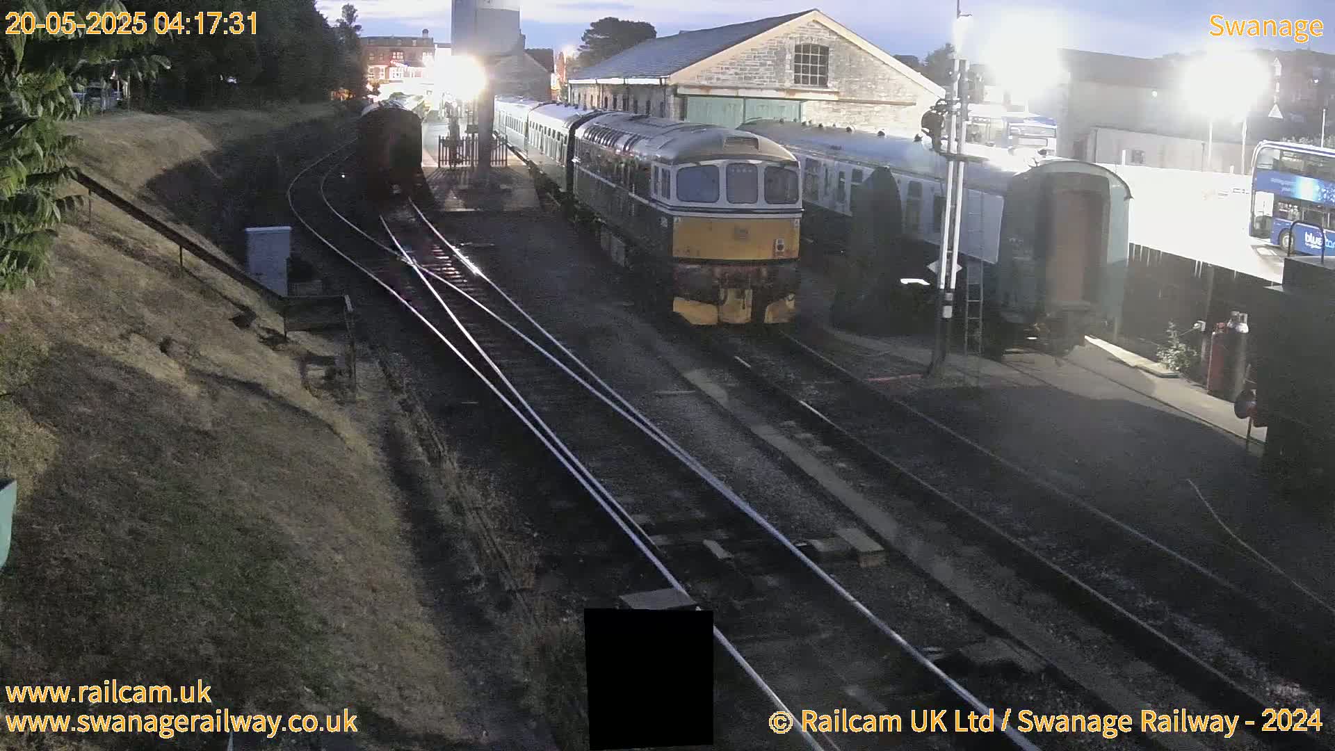 A diesel locomotive and several railway carriages are parked at a train station at twilight, with train tracks extending into a tunnel.