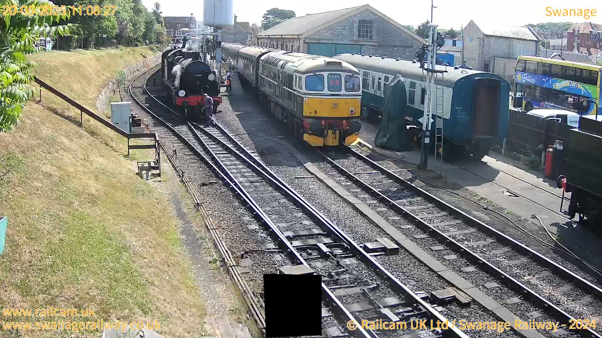 A sunny day at a railway station shows several train cars and locomotives on multiple tracks, with a person standing near one of the locomotives.