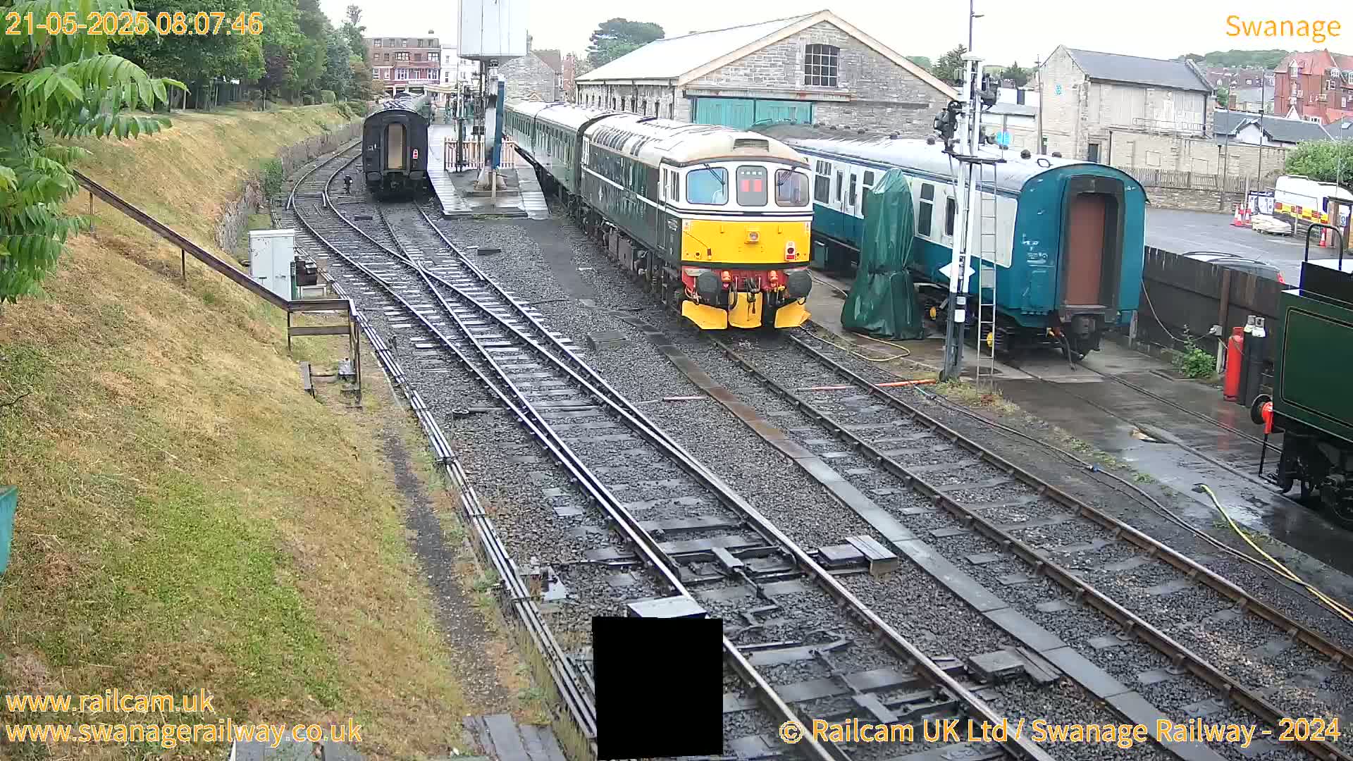 A diesel train is positioned on railway tracks next to several other train cars and a railway station building, on a wet day.