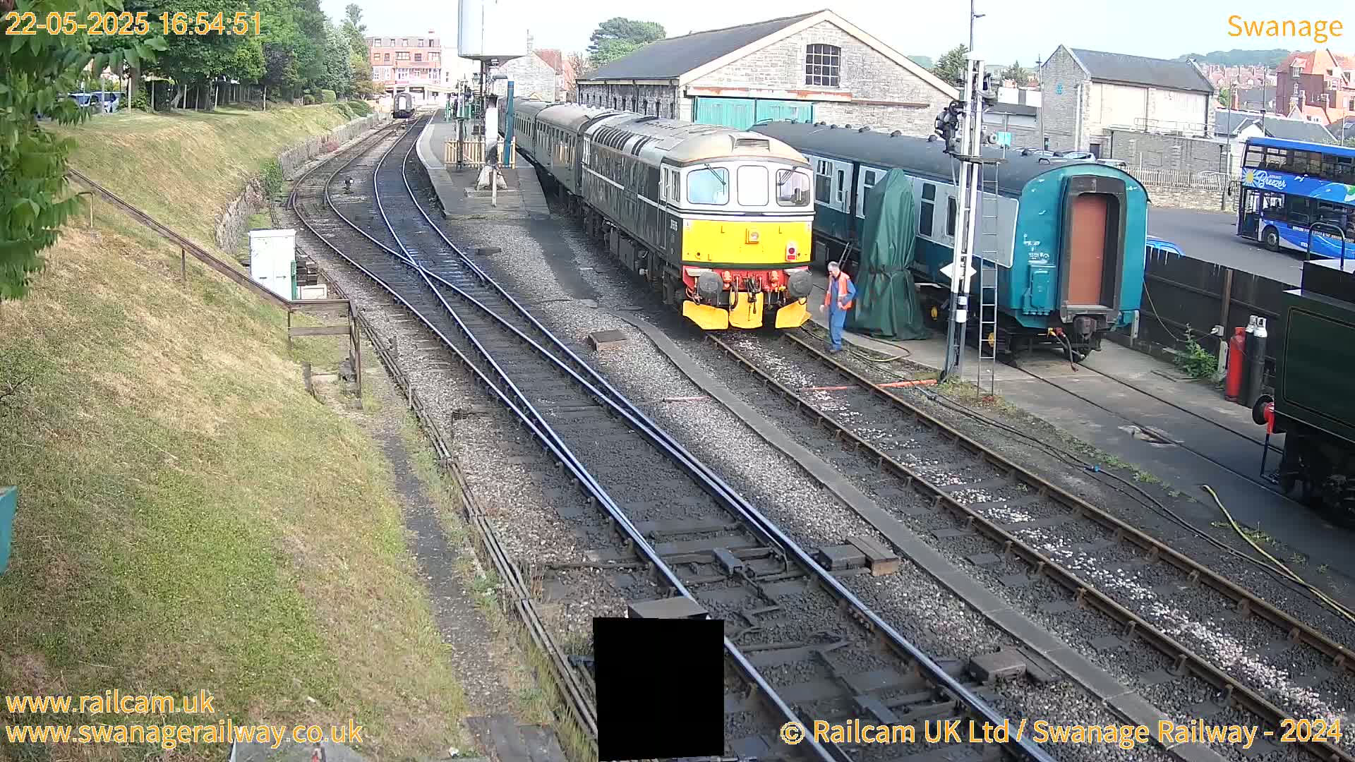 A diesel locomotive and several railway carriages are parked on train tracks at a station on a partly sunny day.