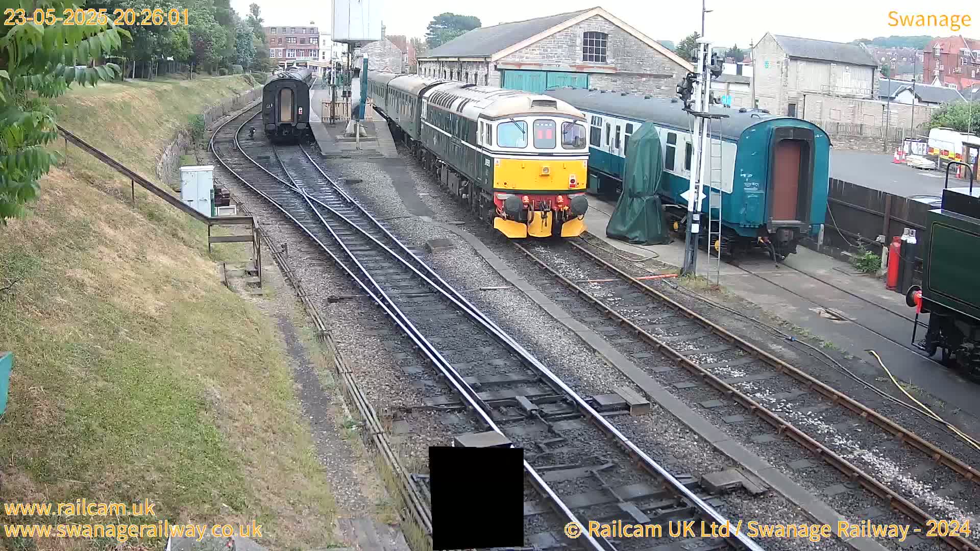 A diesel locomotive leads a train of passenger cars at a railway station on an overcast day.