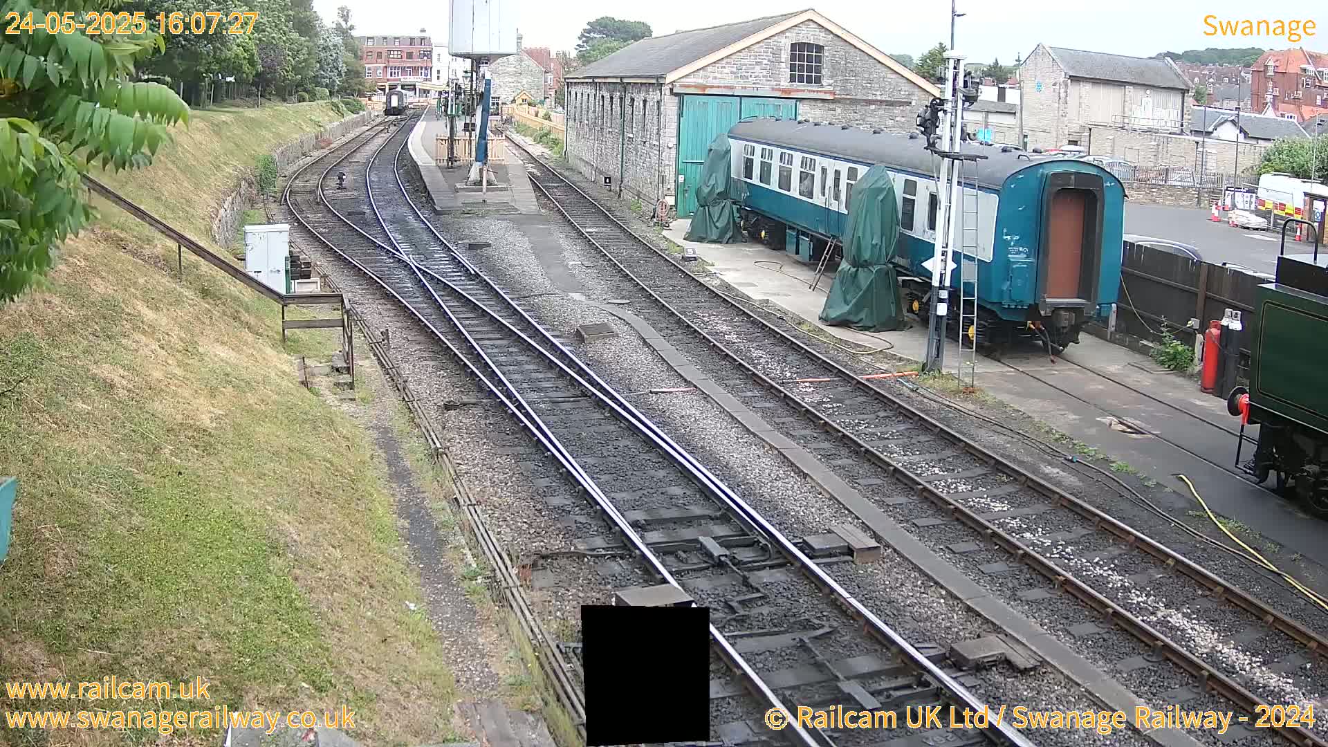 A railway yard with multiple train tracks, a teal railway carriage, and a stone building under an overcast sky.