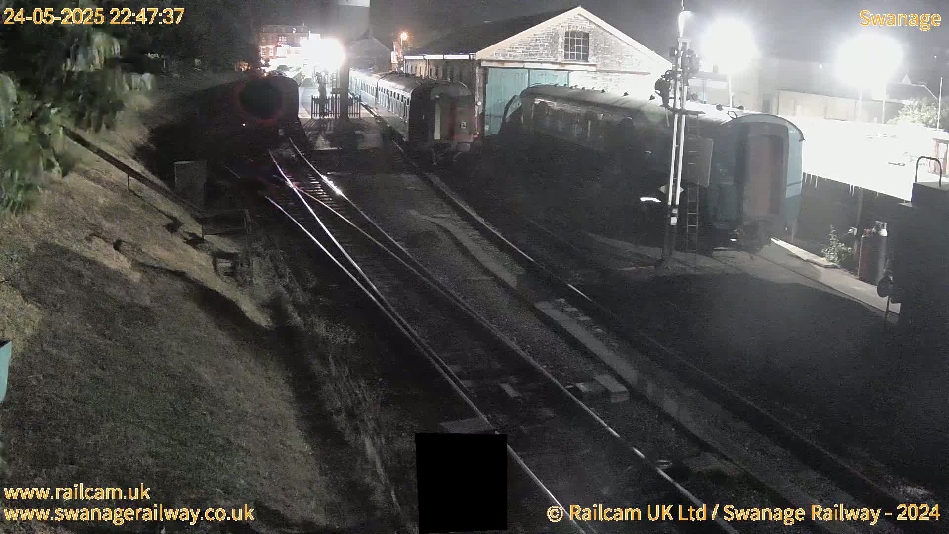 At night, two dark-colored train cars are parked on railway tracks next to a building, with another train car visible further down the tracks.