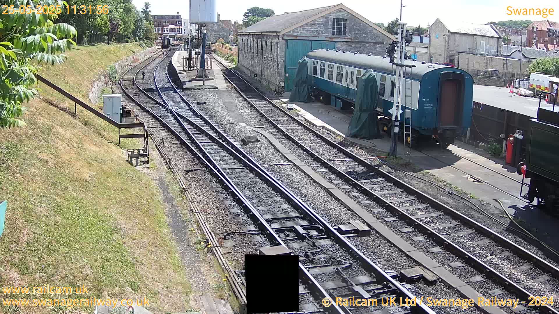 A sunny day at a railway station shows multiple train tracks, a teal railway car parked near a stone building, and grassy areas.