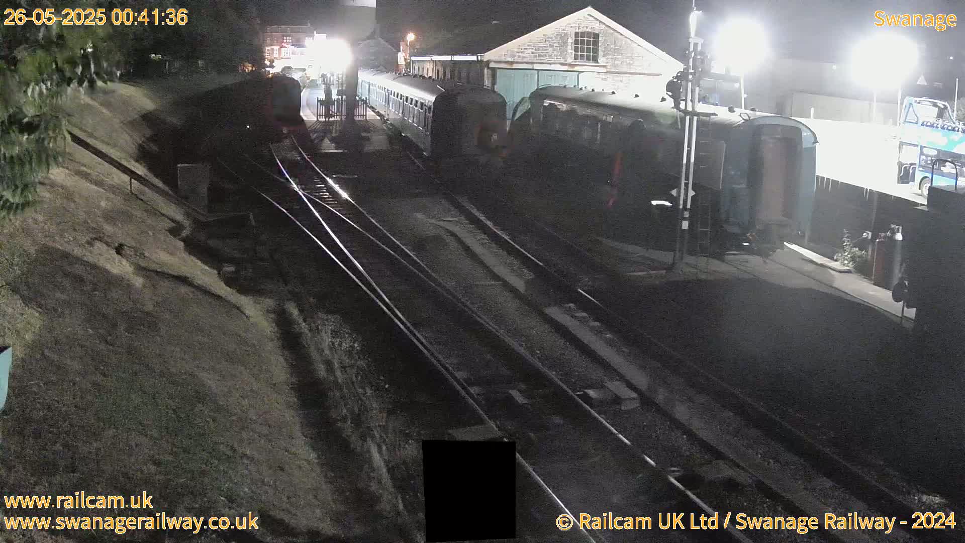 At night, a railway station with several train cars parked on the tracks is seen, illuminated by artificial lighting.