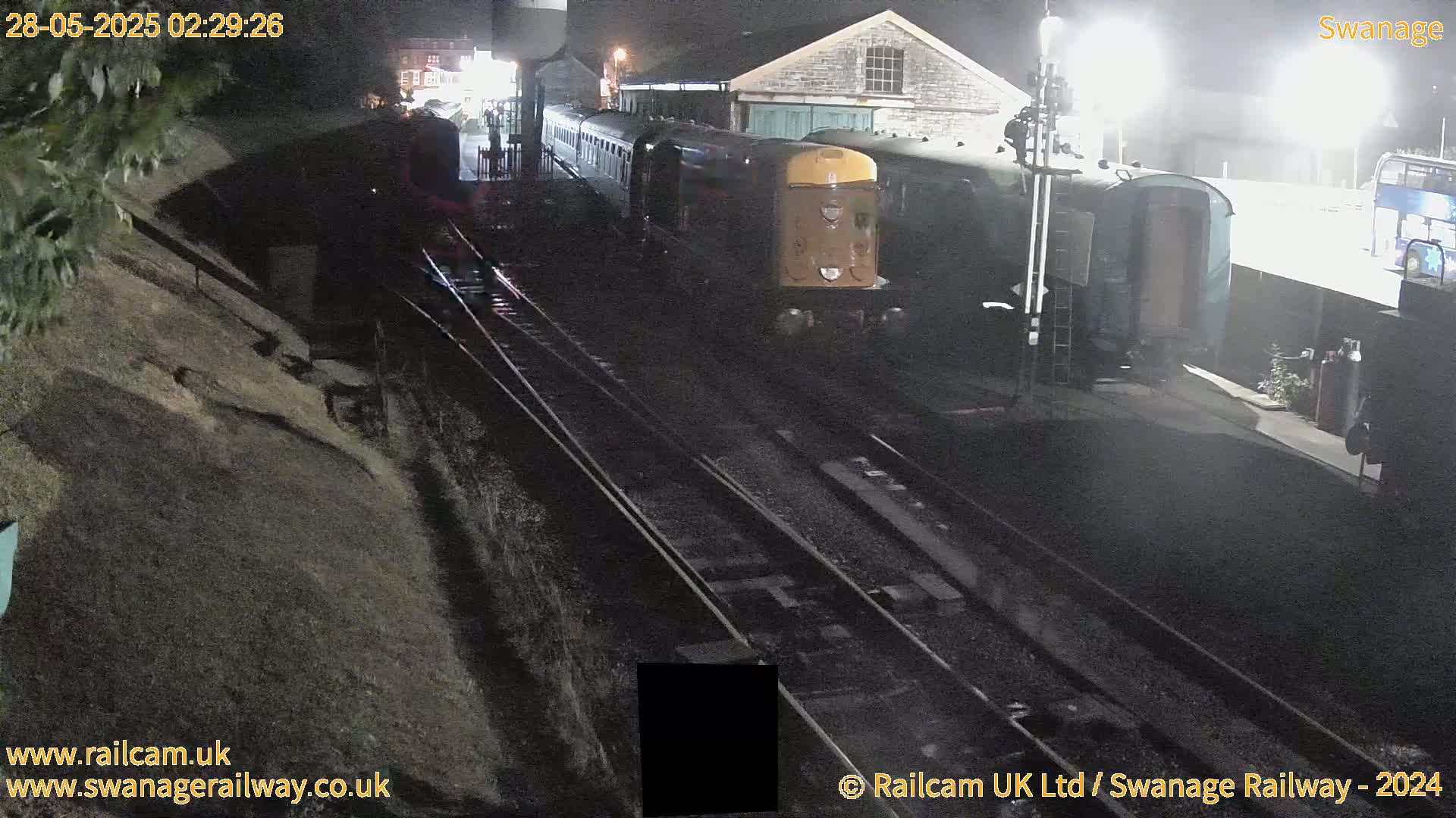 A train is parked at a station at night, next to a building and a hill.