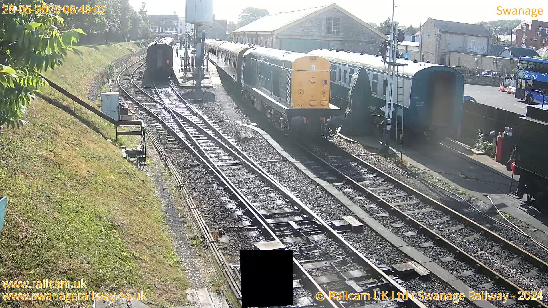 A sunny day at a railway station shows a diesel locomotive and passenger cars parked on multiple tracks, with additional railway infrastructure visible.