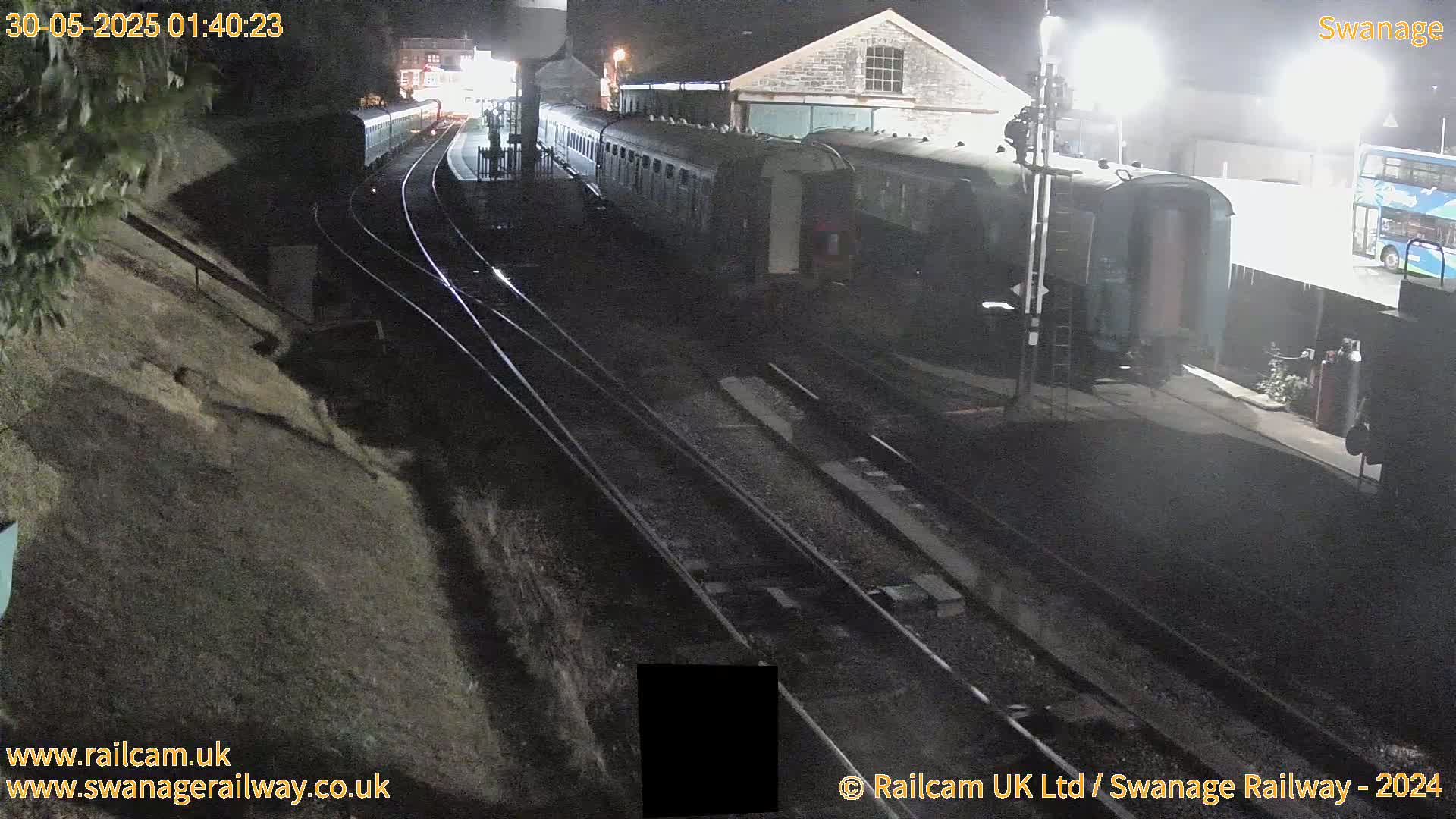 At night, a long train sits on railway tracks next to a building and a bus, with wet tracks and a slightly sloped embankment visible.