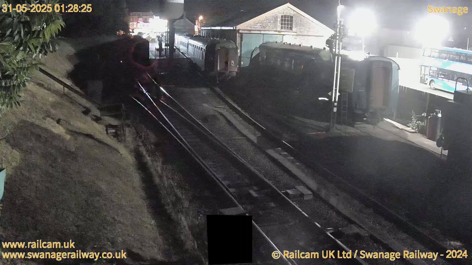At night, several railway carriages are parked at a station next to a bus and a building, with train tracks extending into the dark distance.