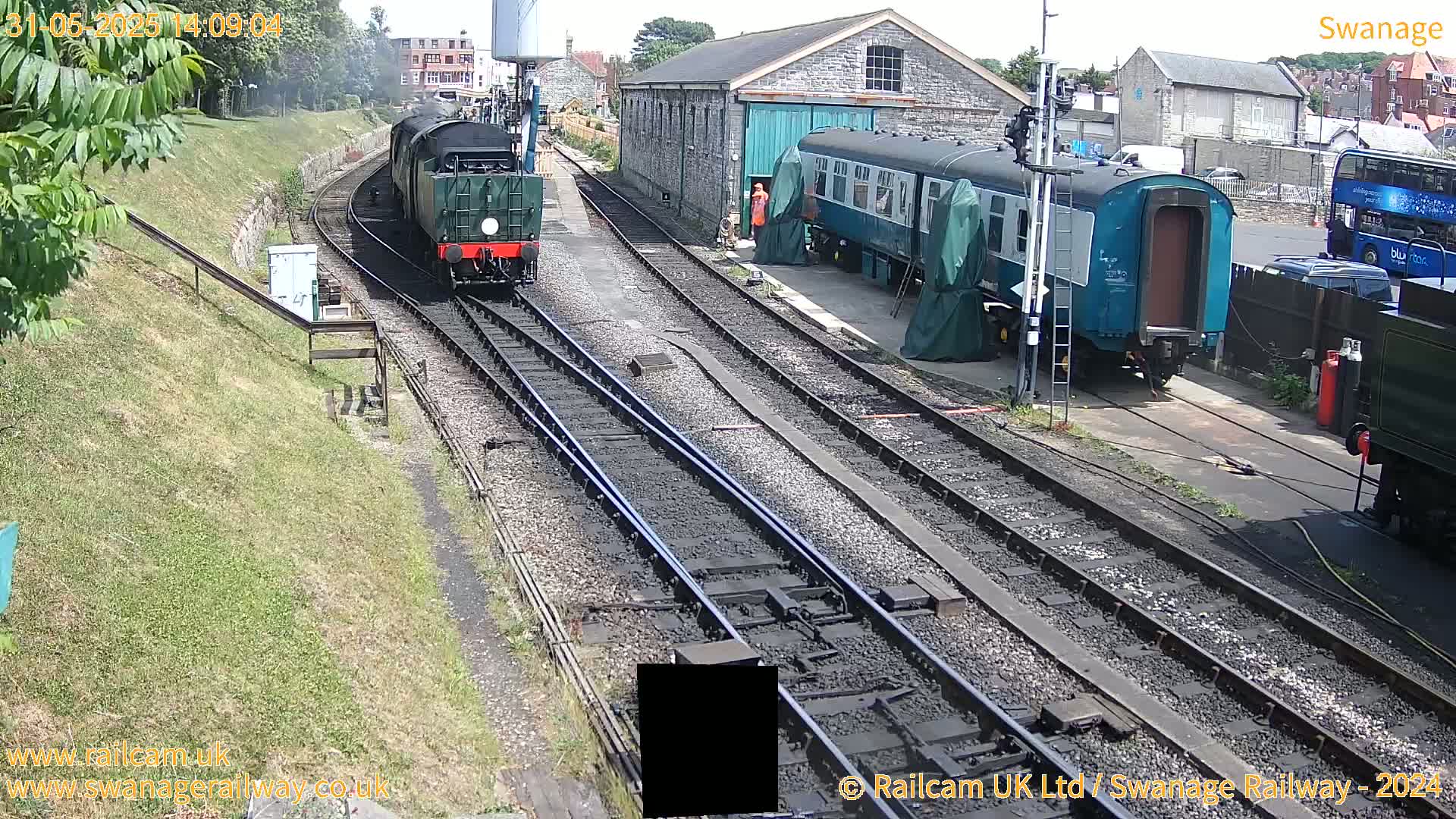 A green steam train is approaching a railway station on a sunny day, where railway carriages are parked beside a stone building.