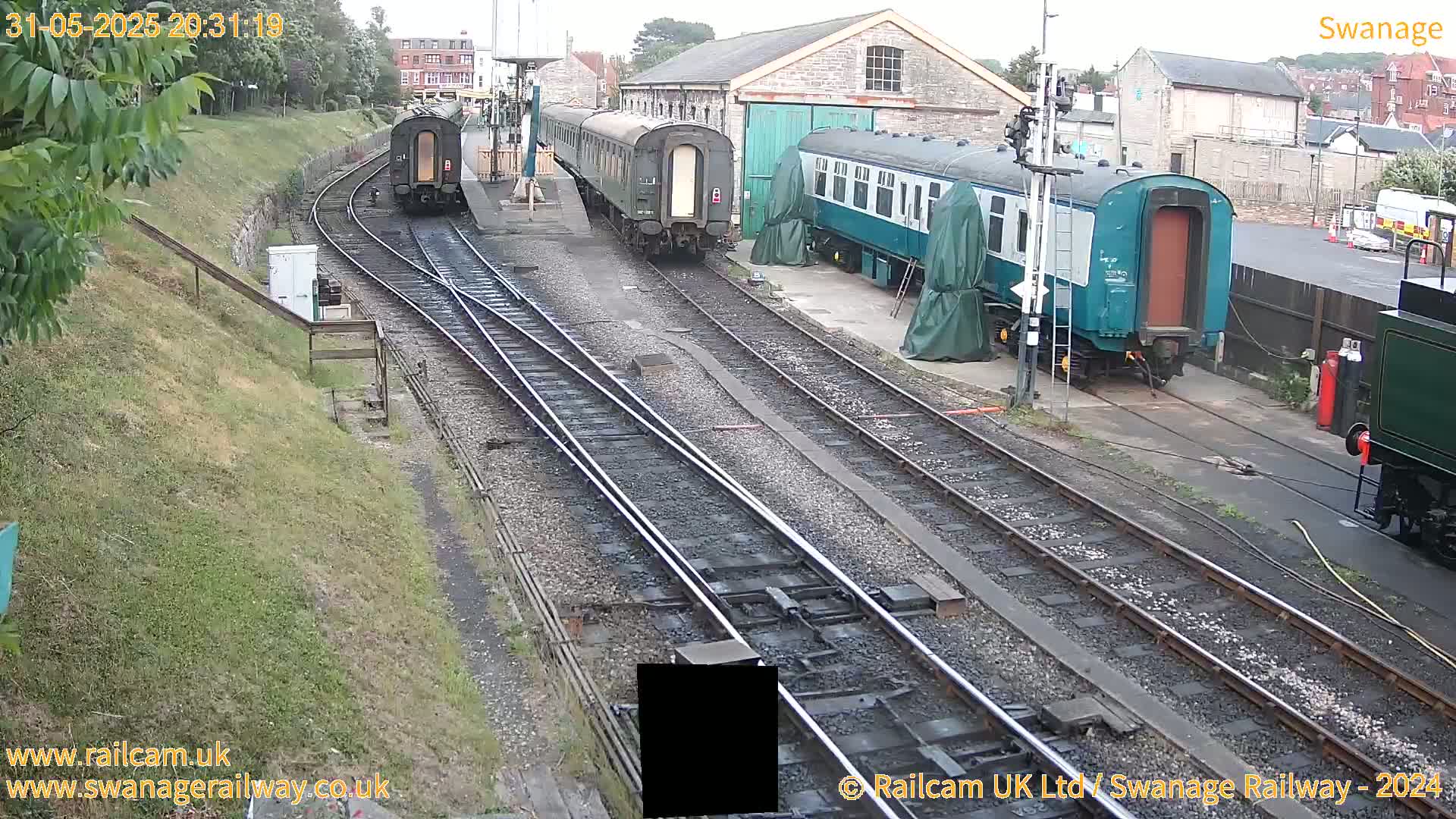 Several railway cars are parked along train tracks next to a railway building, under an overcast sky.