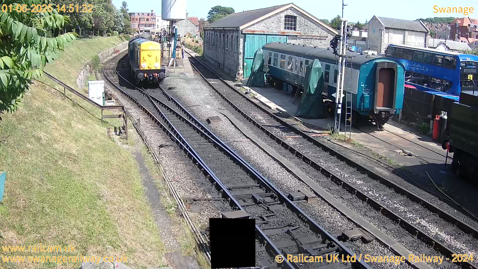 A yellow diesel locomotive is approaching a railway station on a sunny day, with railway carriages and a double-decker bus visible nearby.