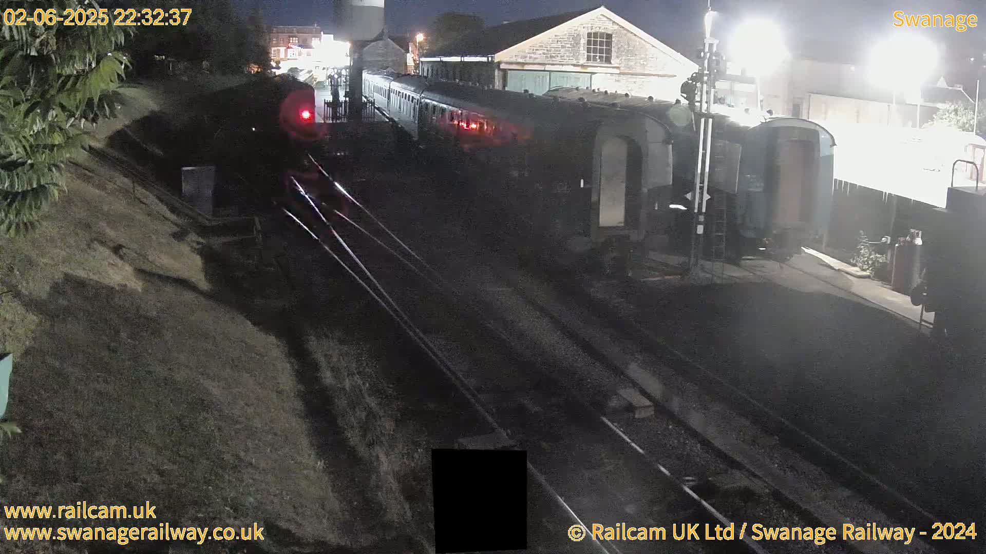 At night, a dark-colored train sits on railway tracks next to a stone building, partially illuminated by artificial lights.