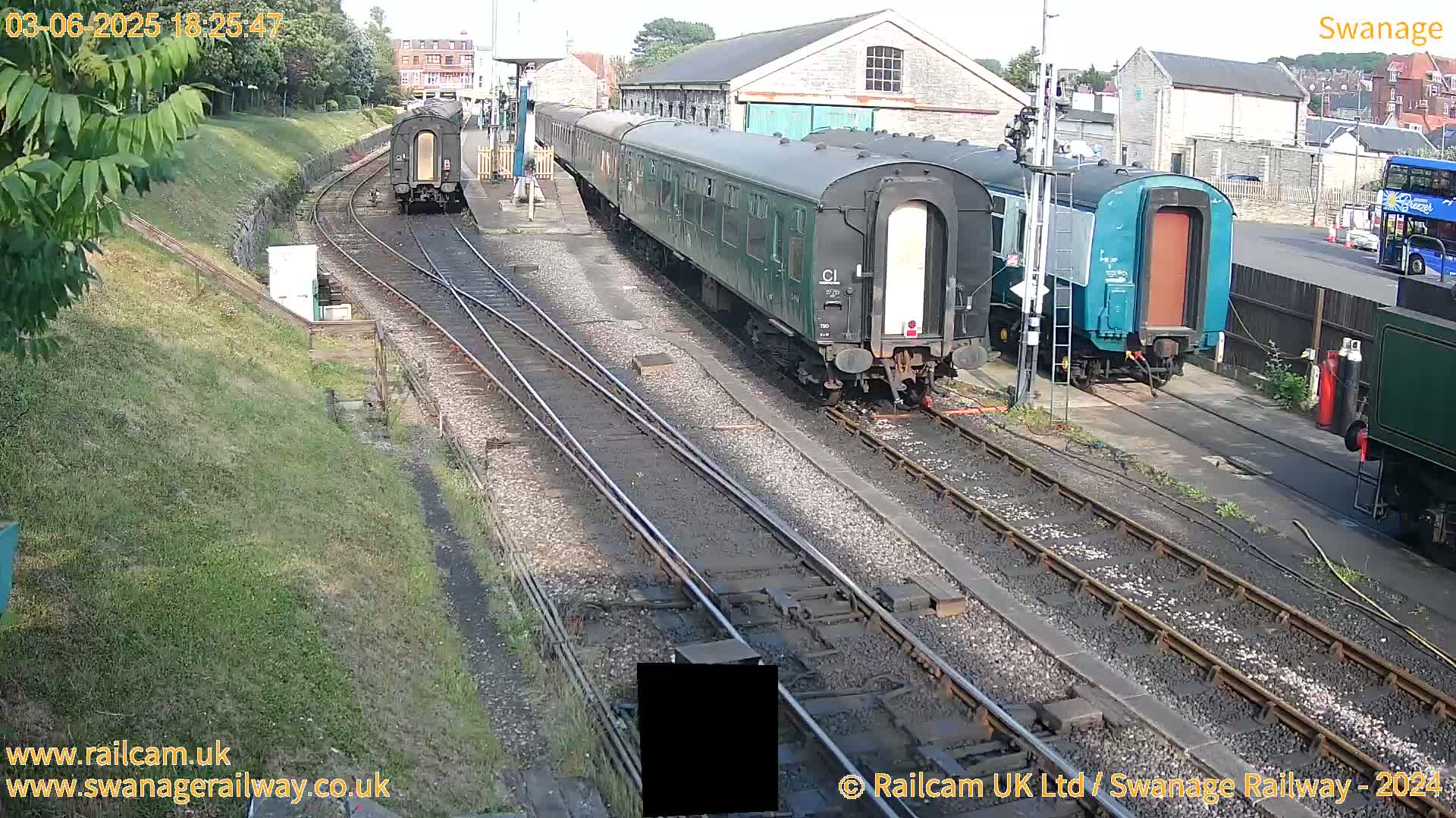 Several railway carriages are parked on train tracks beside a railway station building on a sunny day.