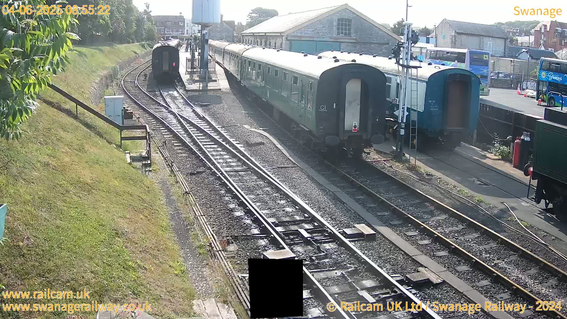Several dark-green passenger train cars are parked on railway tracks next to a building on a sunny day.