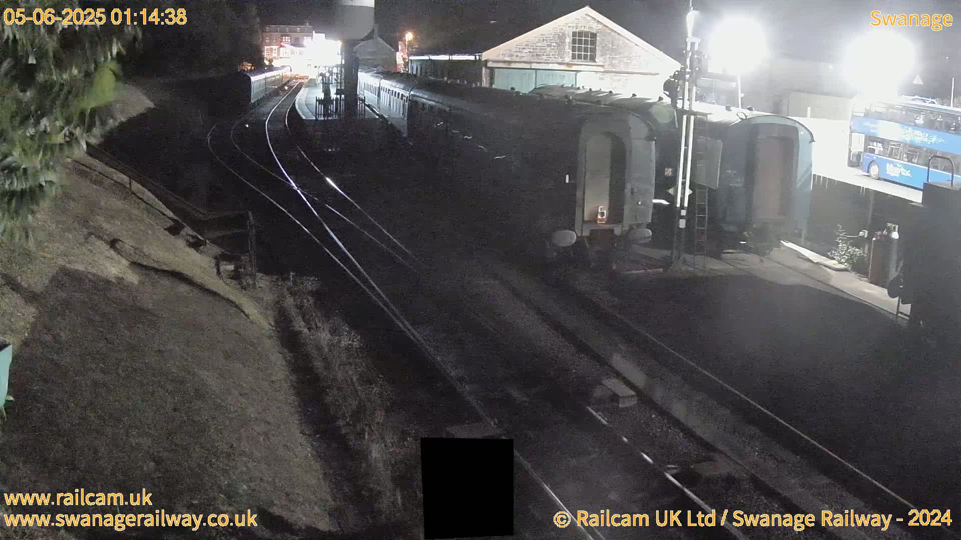 At night, a train sits in a station next to a bus, with train tracks and a sloped embankment visible.