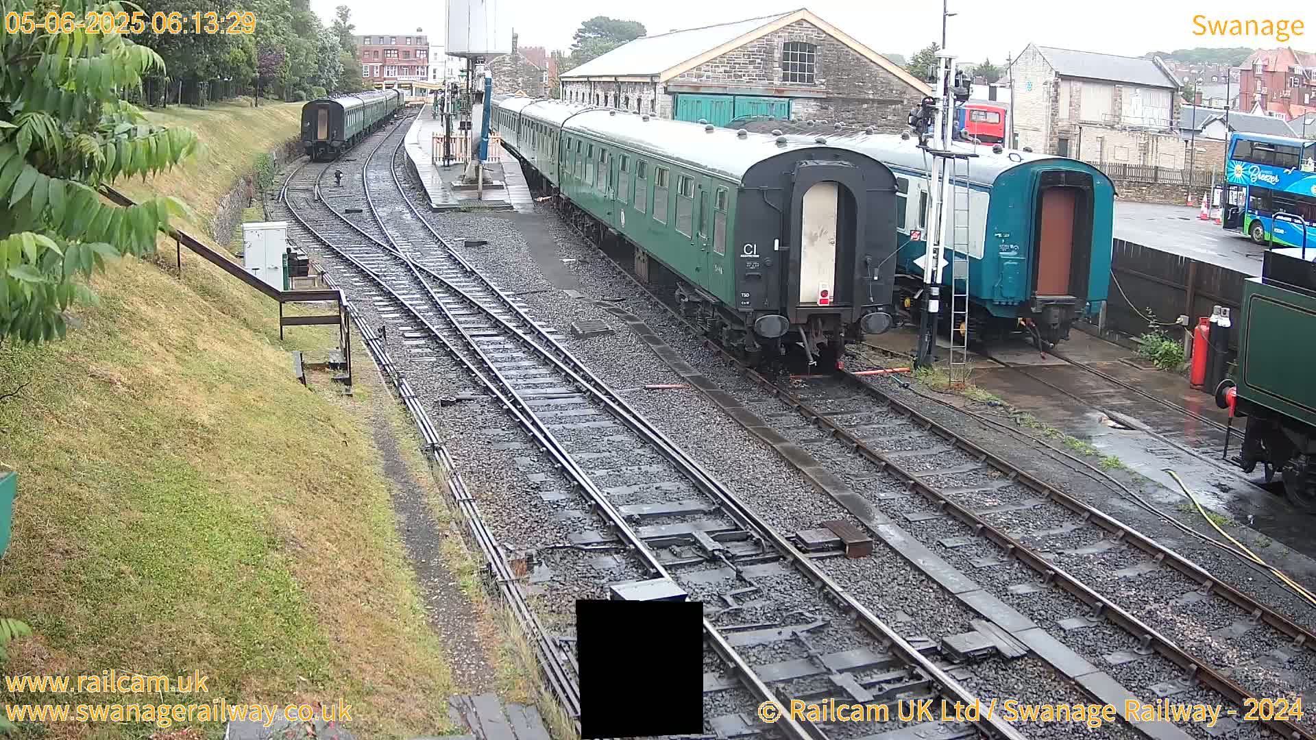 Several dark-green railway carriages are parked on rain-slicked train tracks next to a railway station building.