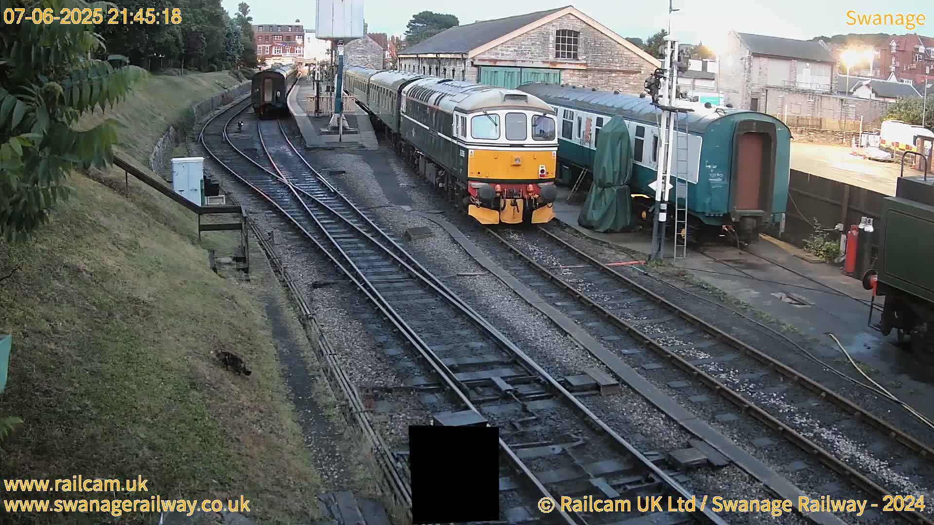 A diesel locomotive and several passenger cars are parked on train tracks at a station under a twilight sky.
