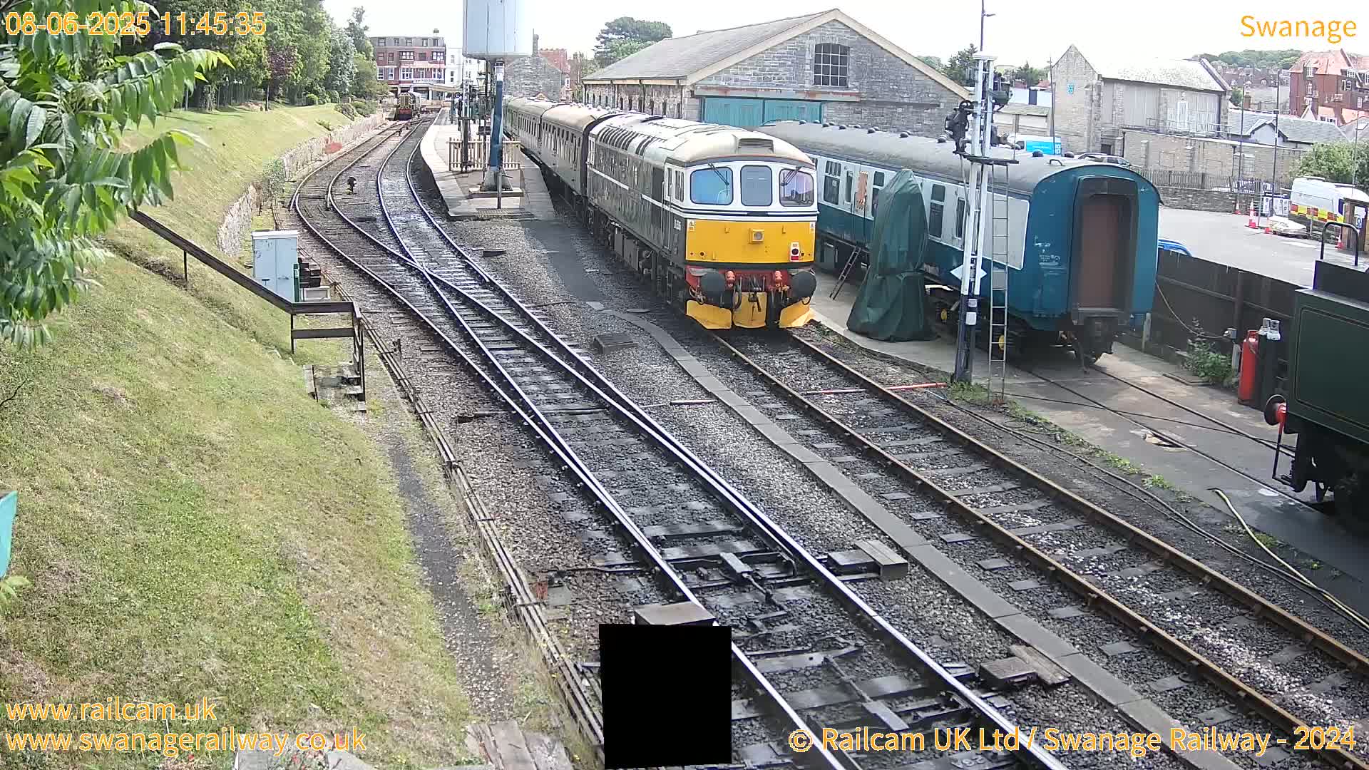 A diesel locomotive pulls a train of passenger cars on railway tracks next to a railway station building on a partly sunny day.