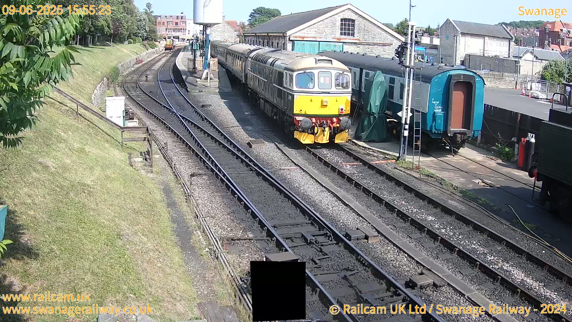 A yellow and dark-green diesel locomotive leads a train of passenger cars at a railway station on a sunny day.