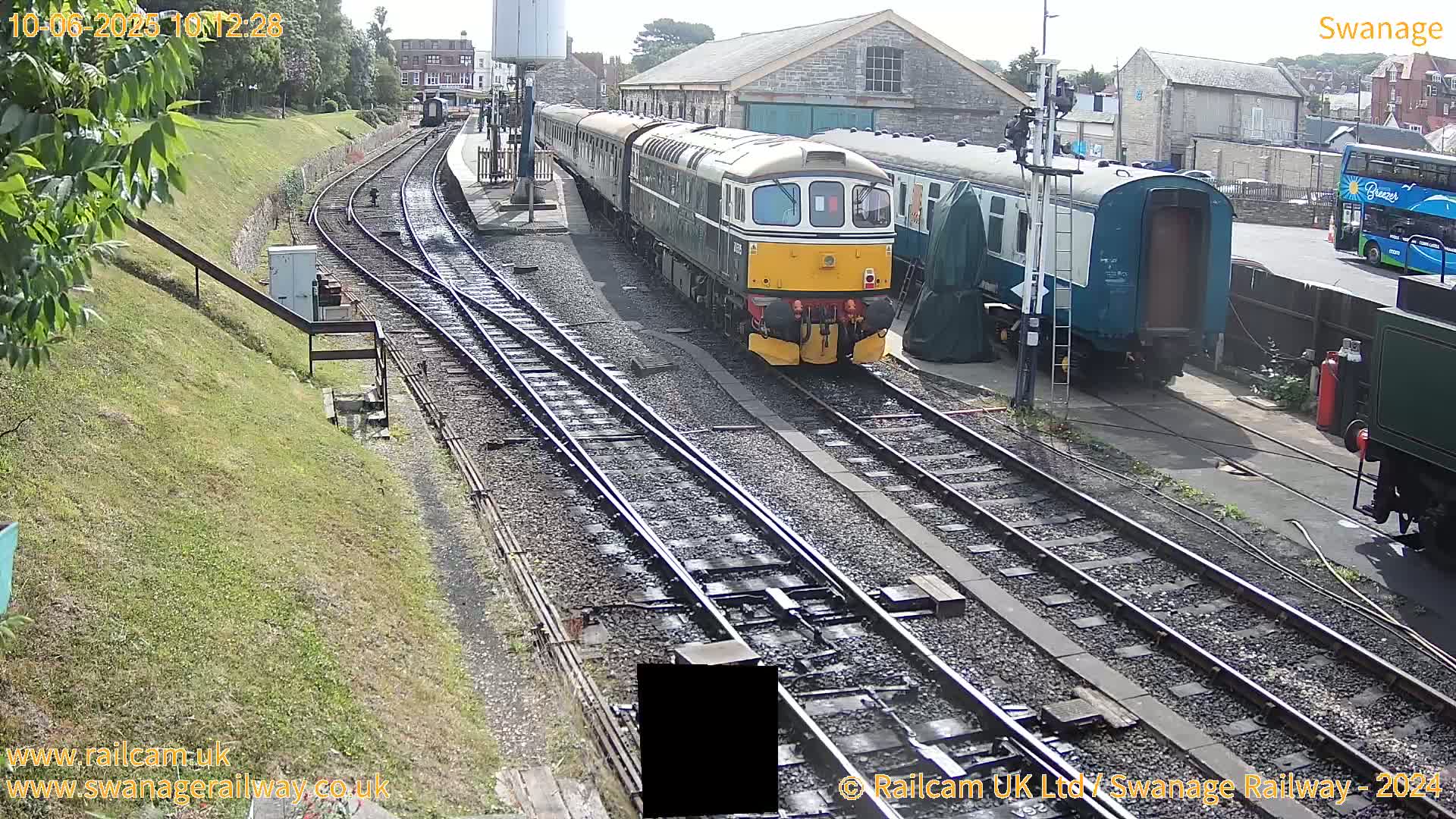 A diesel train pulls into a railway station on a sunny day, with other train cars and a bus visible nearby.