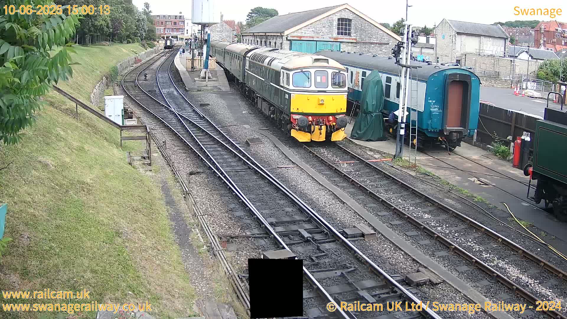 A diesel locomotive and several railway carriages are parked on train tracks next to a railway station building on a partly sunny day.