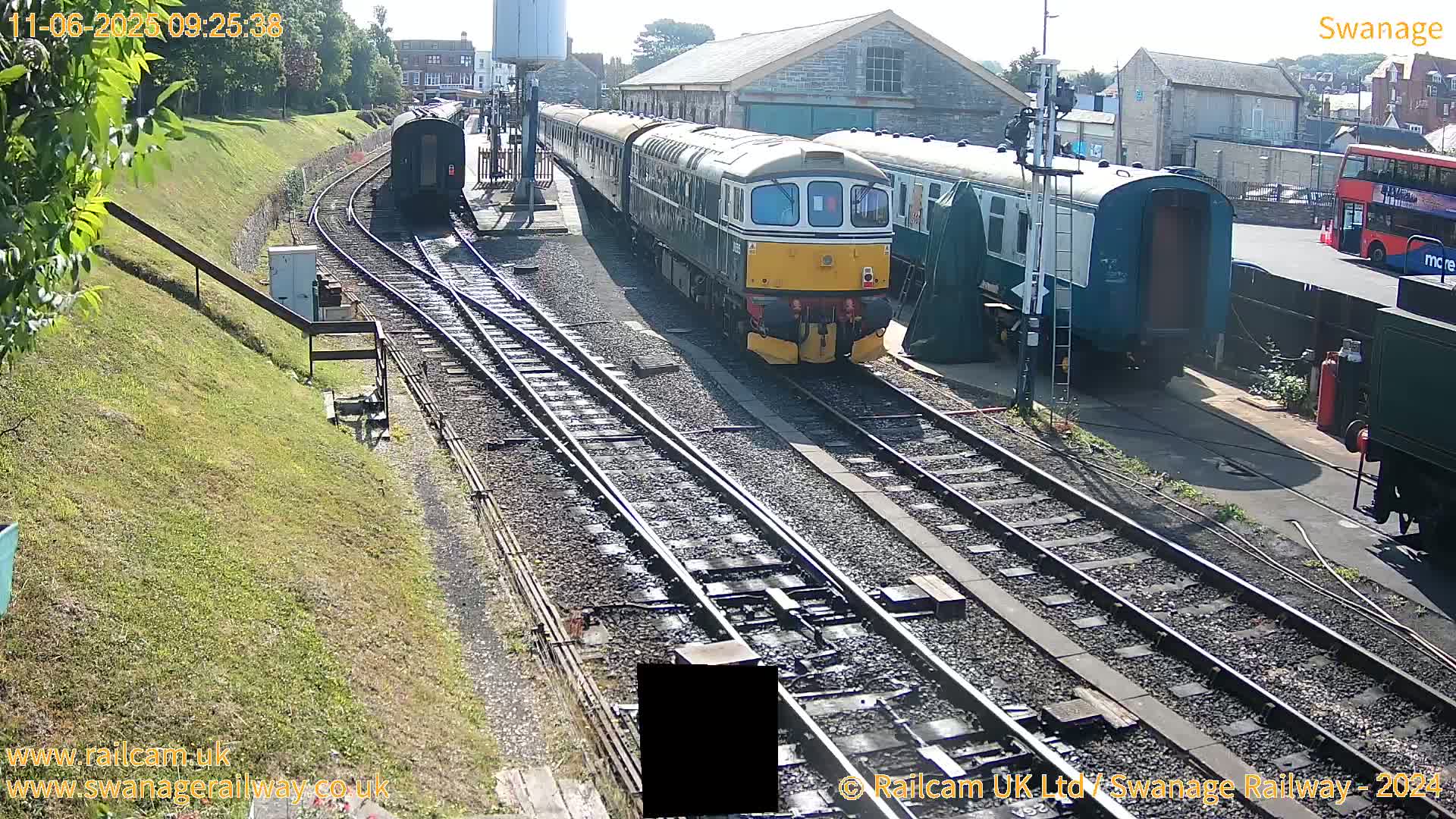 A diesel train sits on railway tracks next to several other train cars and buildings on a sunny day.