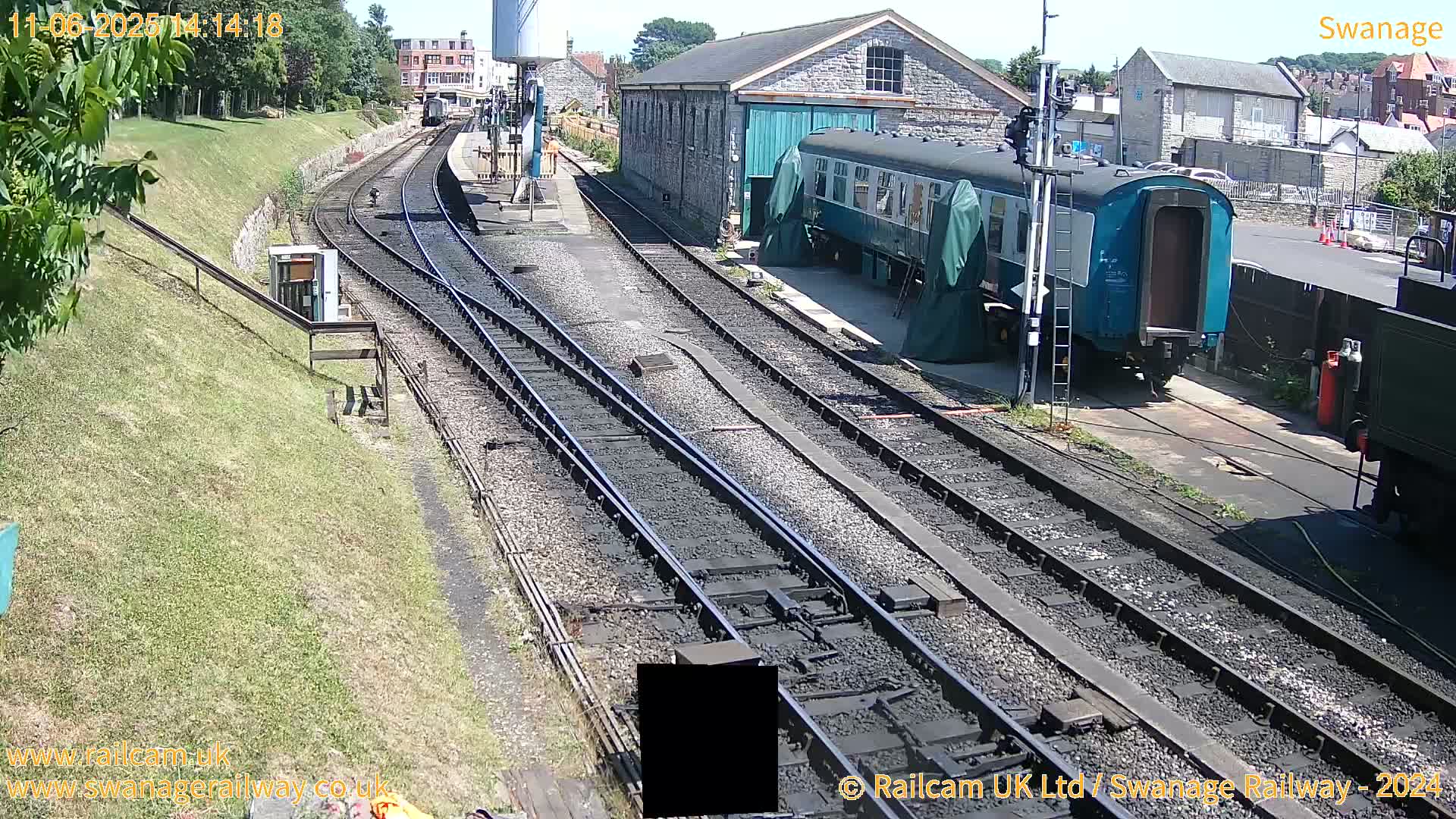A sunny day at a railway station shows multiple train tracks, a train car partially covered, and a stone building; grassy embankments are visible on the left.