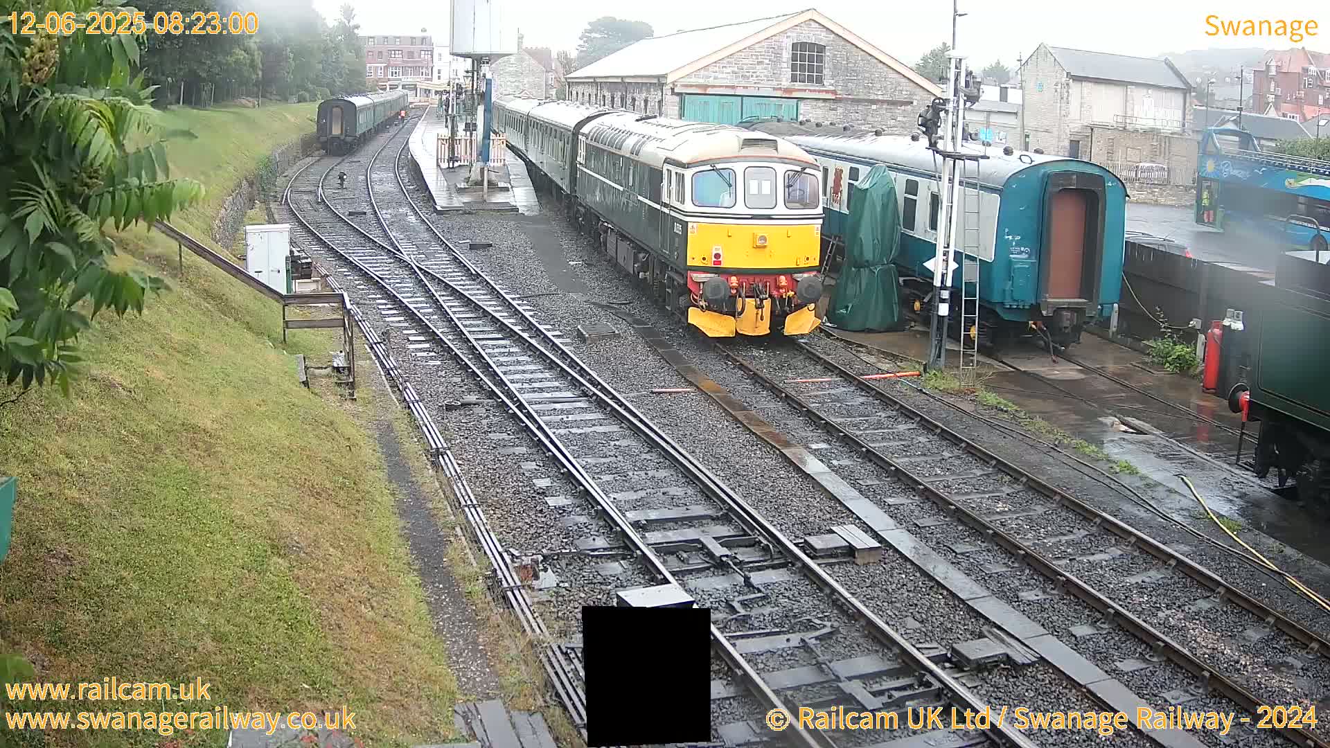A diesel locomotive and several railway carriages are parked on wet railway tracks next to a stone building in rainy weather.