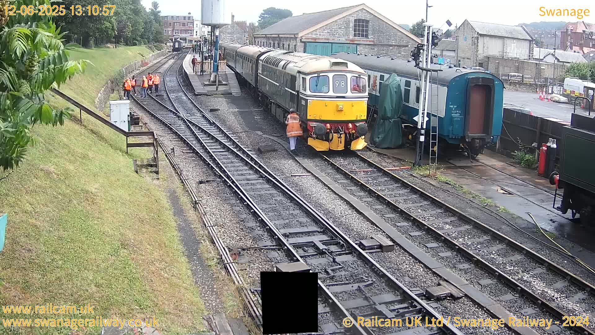 A diesel locomotive and passenger train cars are at a railway station on a wet day, with several people in orange high-visibility vests standing nearby.