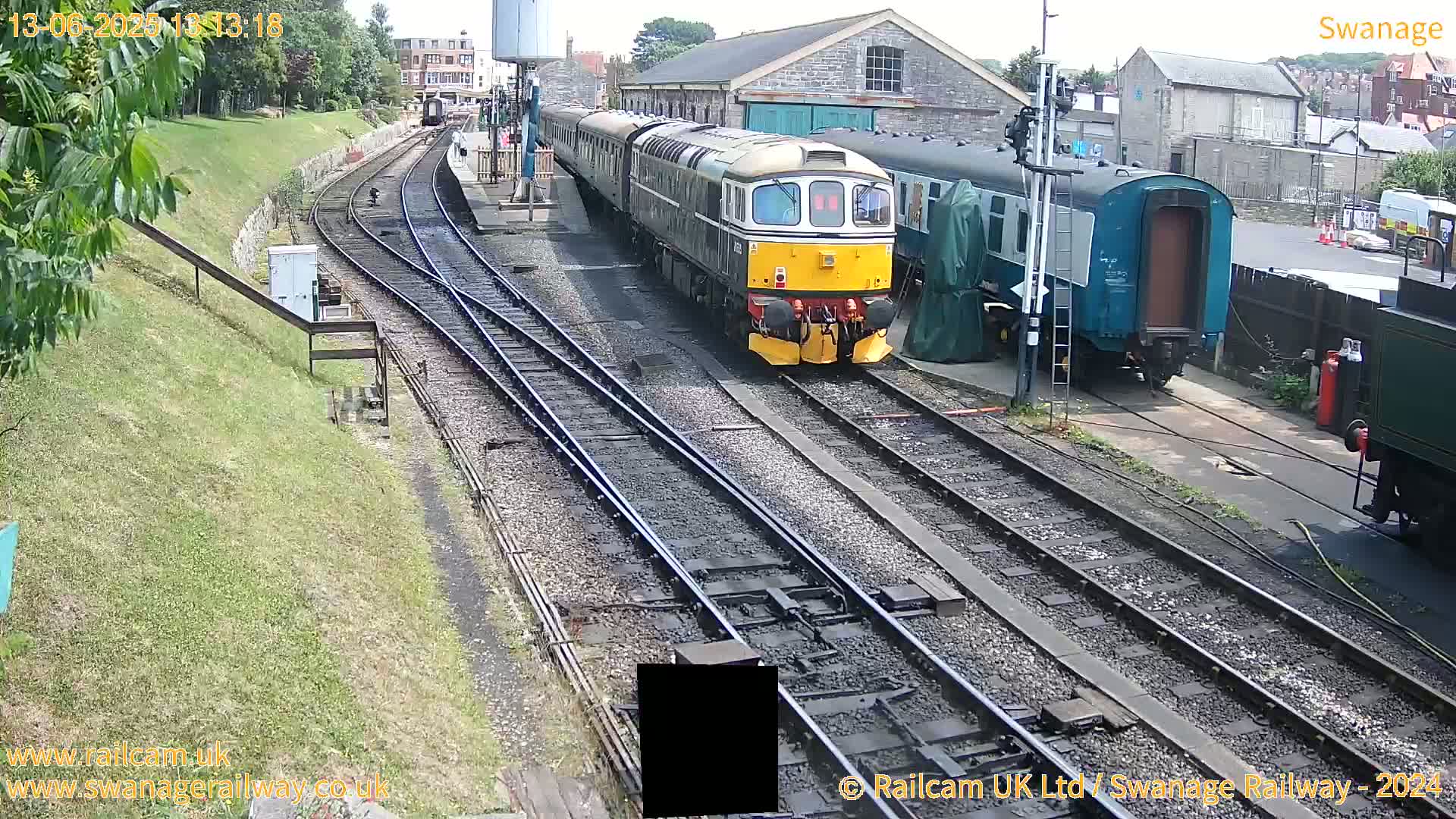A yellow and black diesel train is arriving at a railway station on a sunny day, with other train cars and buildings visible nearby.