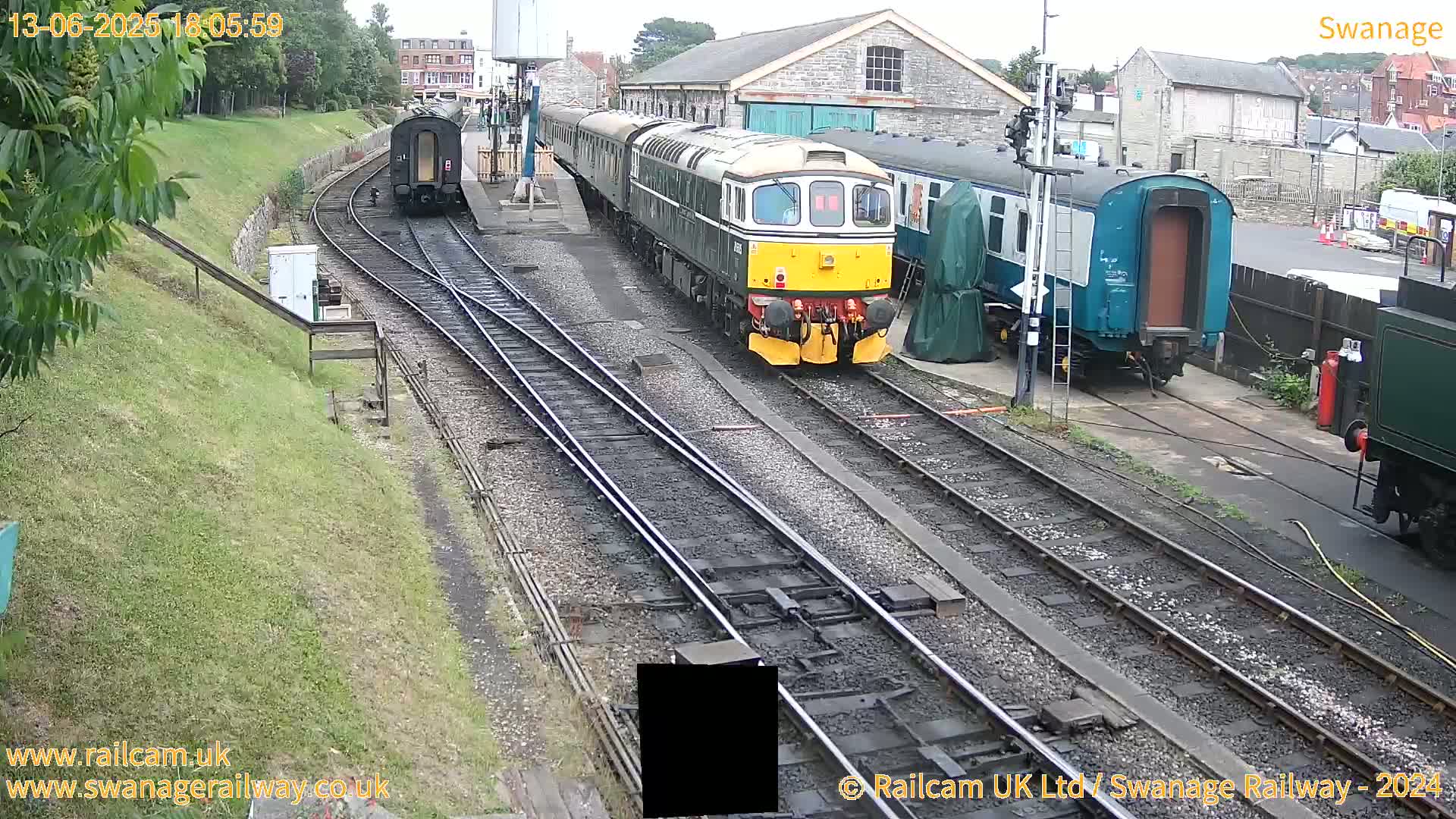 A diesel locomotive and several passenger cars sit on railway tracks in front of a railway station building on an overcast day.