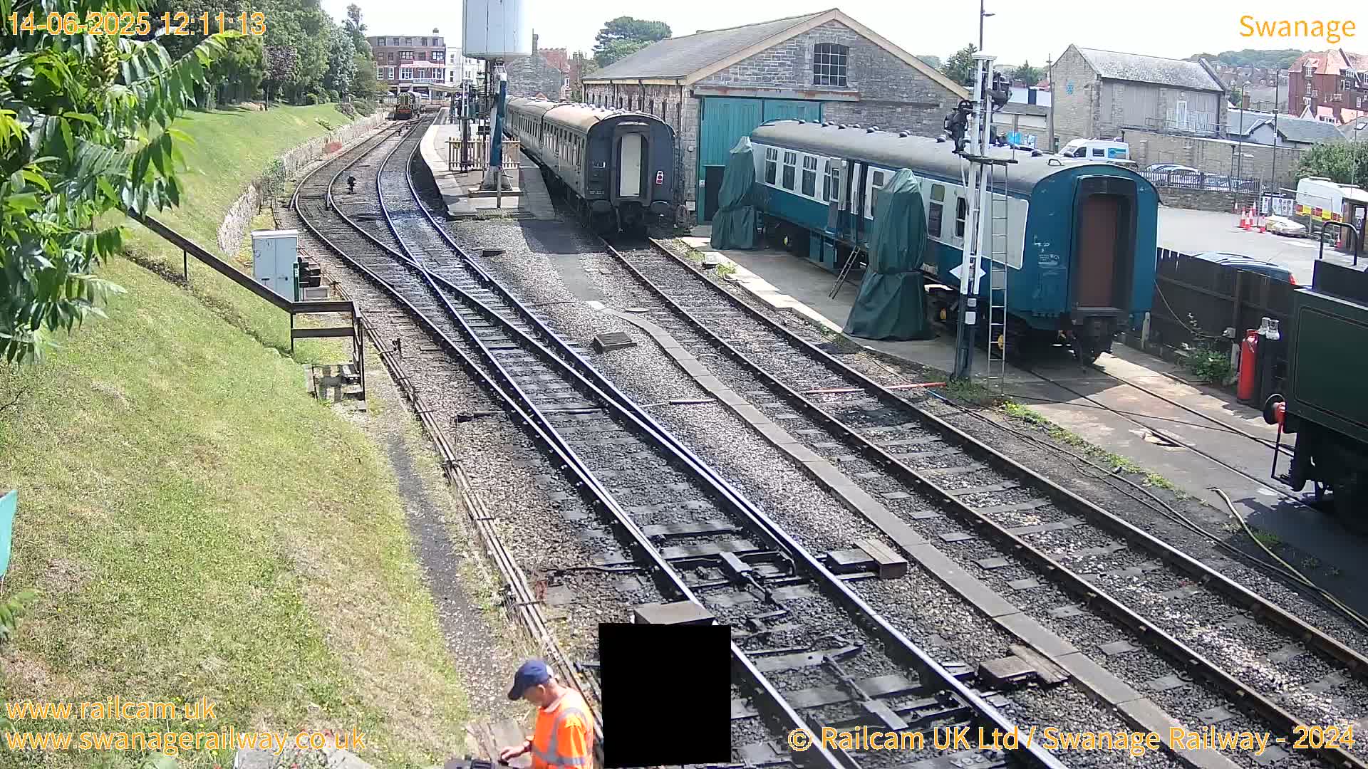 A sunny day at a railway station shows several train cars parked alongside multiple train tracks, with a person in a high-visibility vest near the tracks.