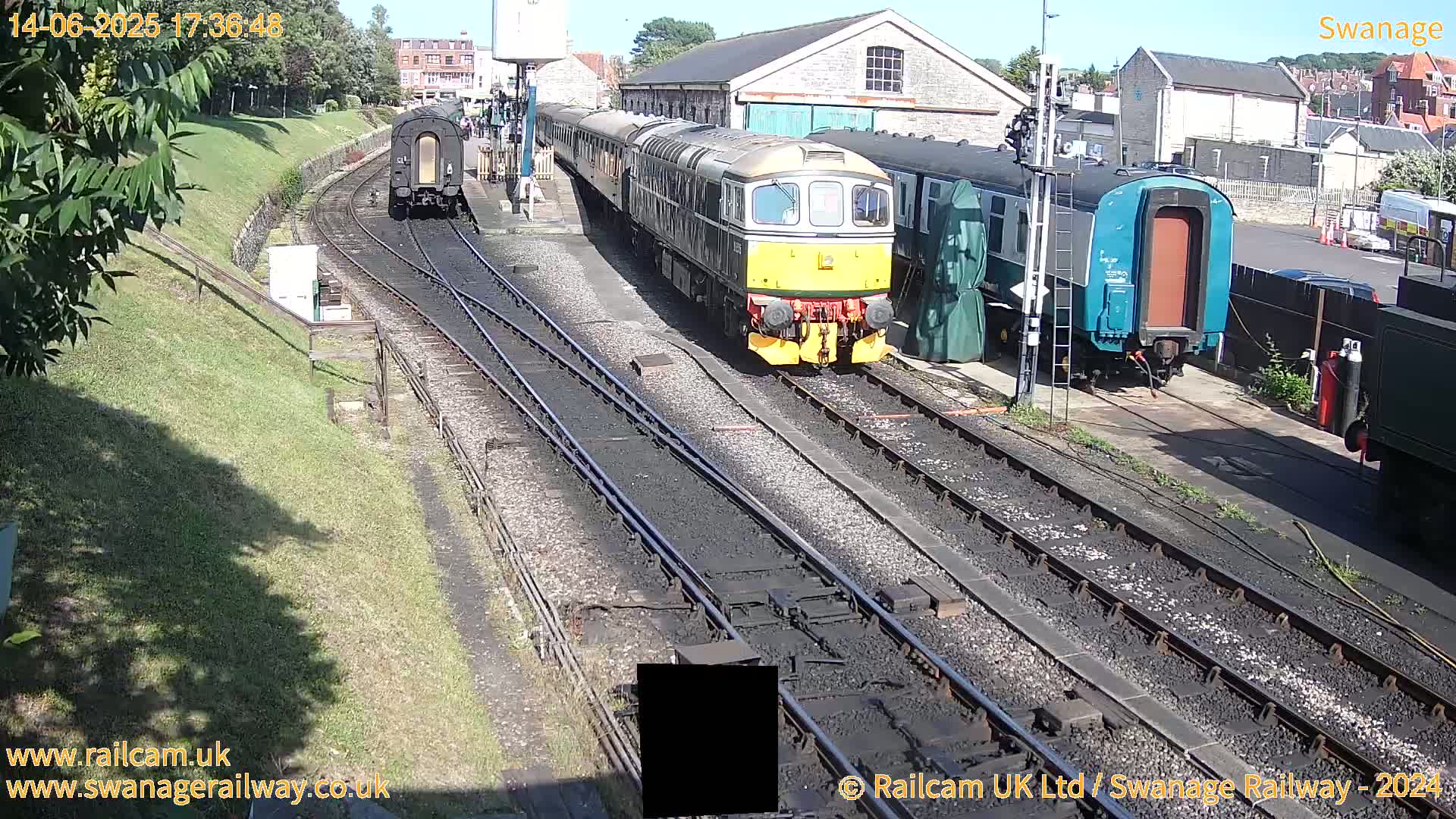 A diesel locomotive leads a passenger train past a rail yard on a sunny day, with other rail cars visible in the background.
