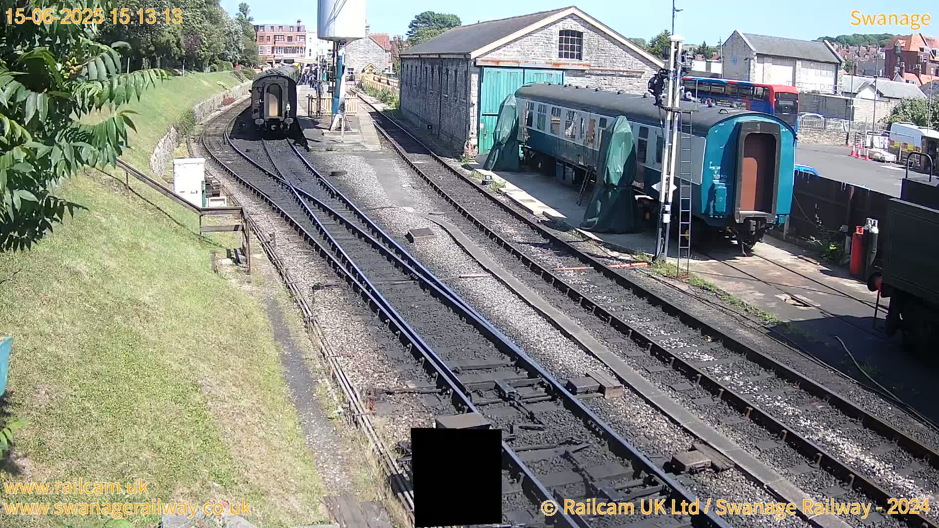 A sunny day at a train station shows railway tracks, several train cars, and stone buildings.