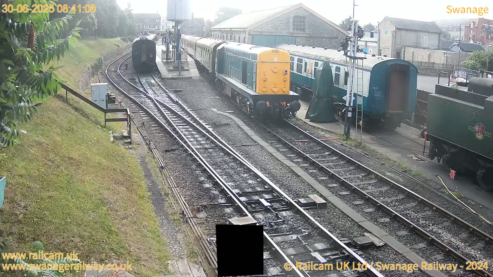 A diesel locomotive pulls a passenger train past a railway station on an overcast day.