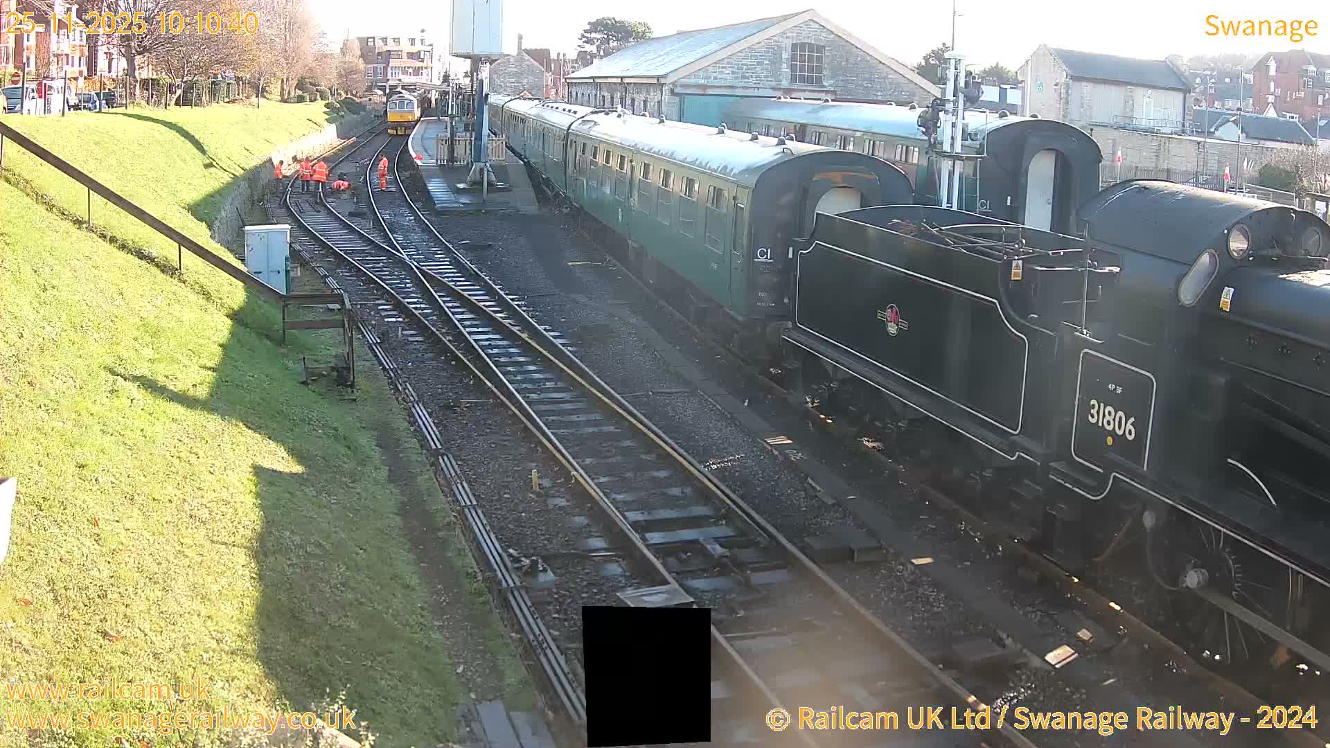 A dark green steam locomotive (31806) and passenger carriages are at a railway station on a clear, sunny day, with several workers in orange high-visibility clothing on the tracks and another yellow train visible in the distance.