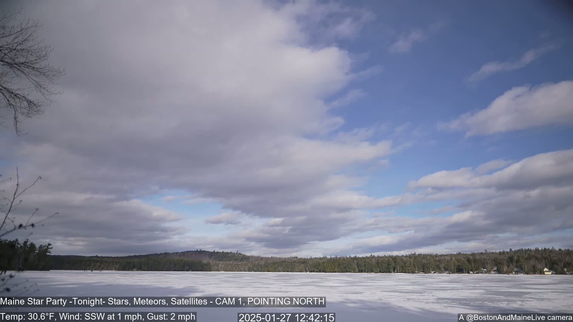 A partly cloudy sky over a frozen lake and a snow-covered shoreline with a background of dark, distant trees.
