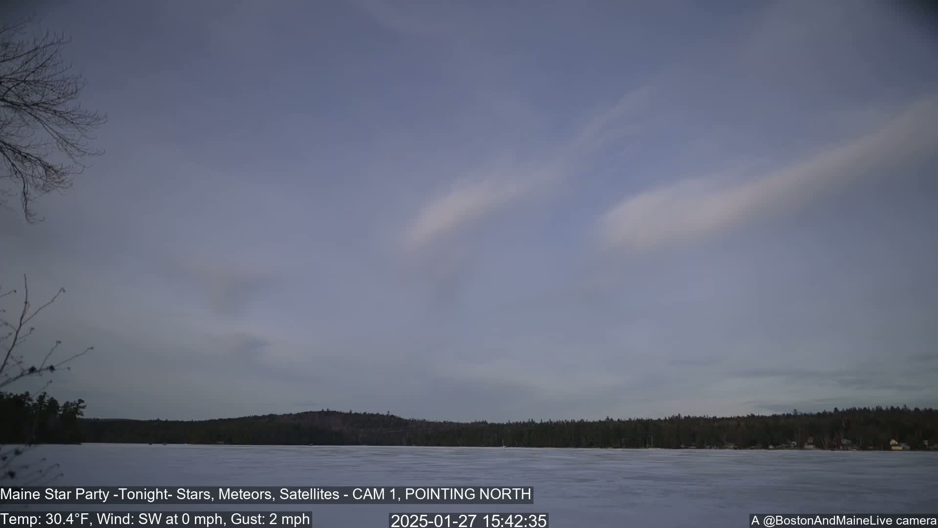 A frozen lake is visible beneath a mostly clear, light-grey sky with wispy clouds, and a dark treeline is in the background.