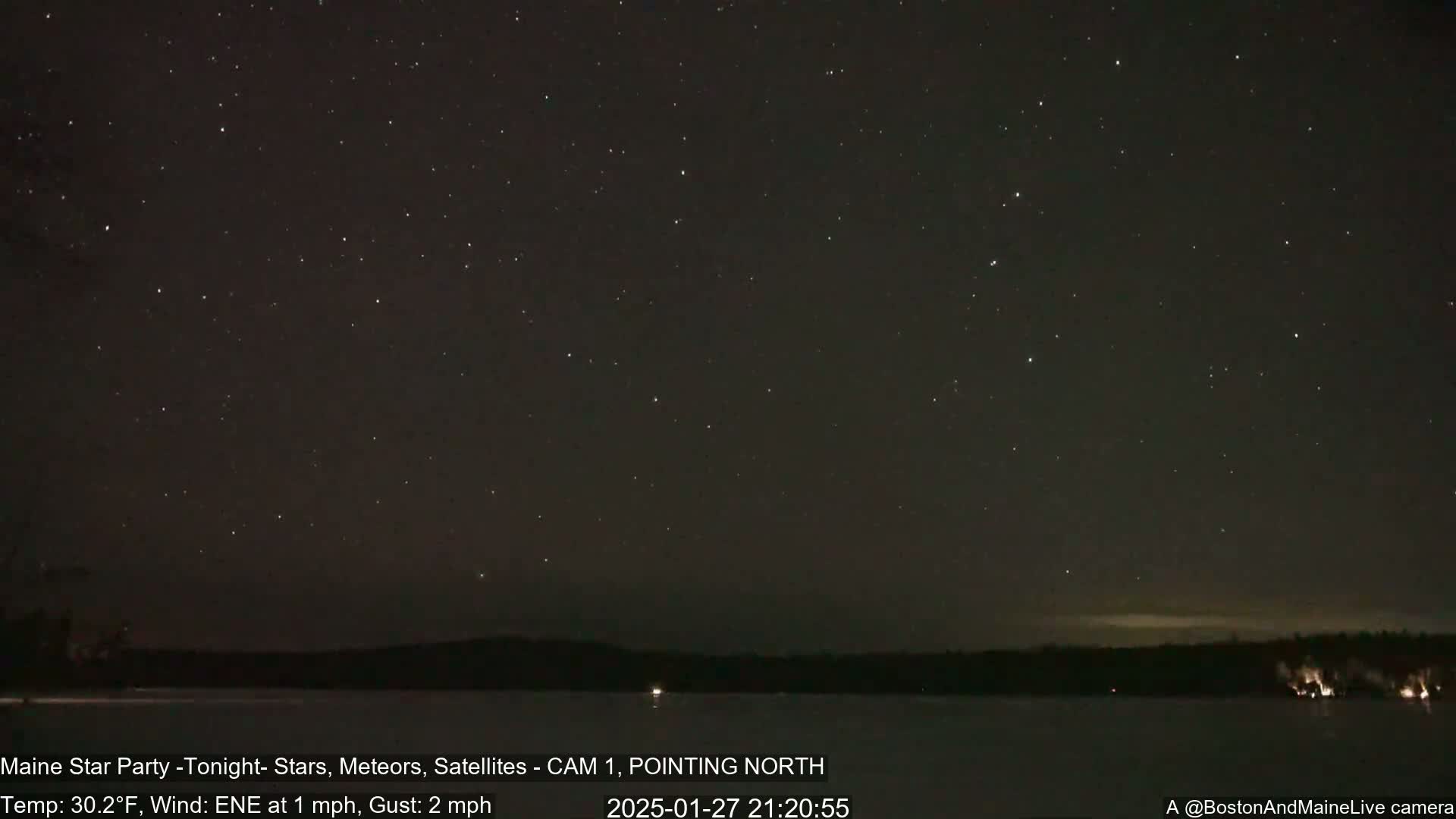 A dark, clear night sky filled with stars is visible above a calm body of water and a distant tree line, with a gentle east-northeast wind.