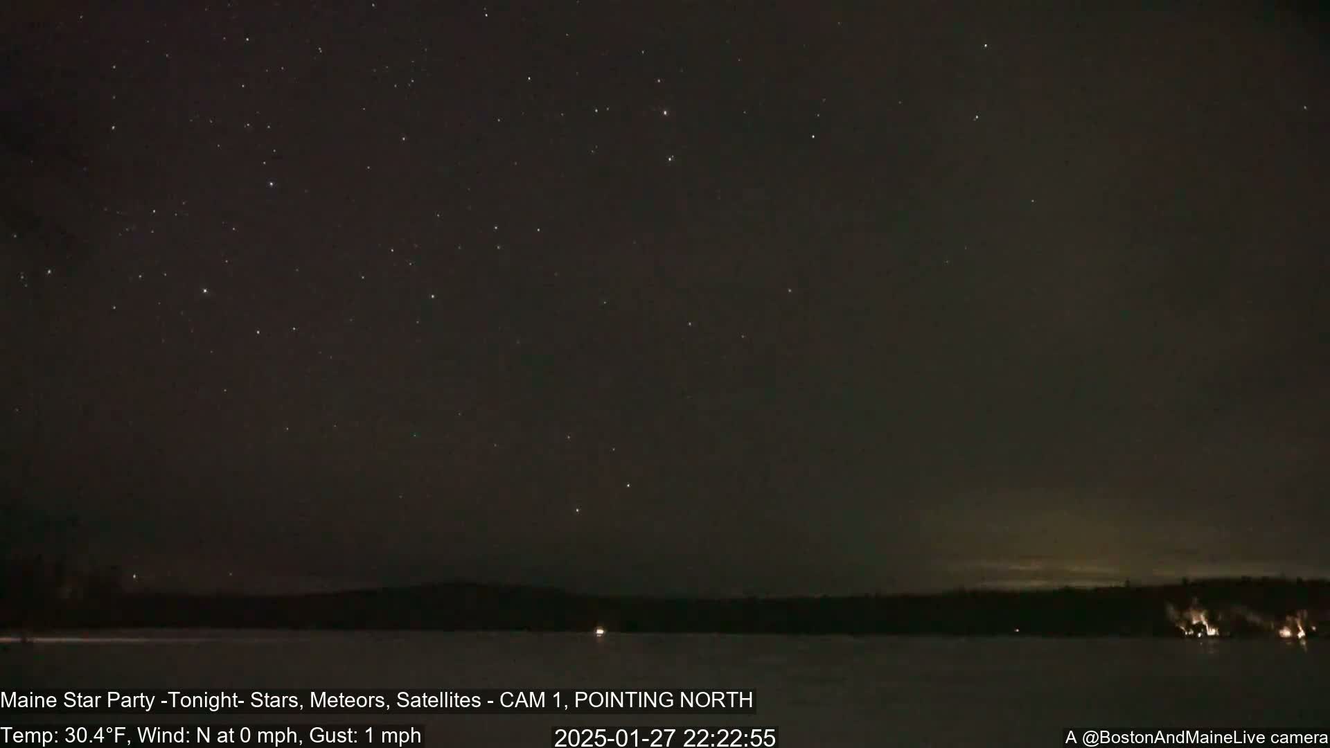 A clear, cold night sky filled with stars and a dark, still body of water with a treeline in the distance.