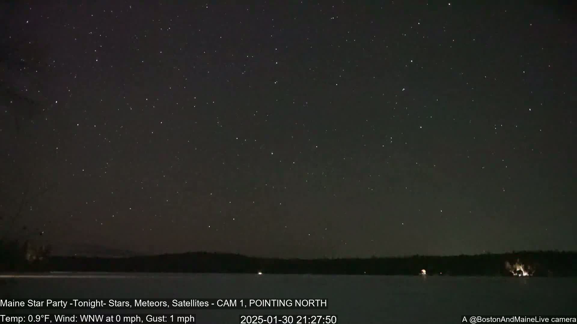 A nighttime view of a star-filled sky over a dark, frozen lake and treeline under calm, cold conditions.