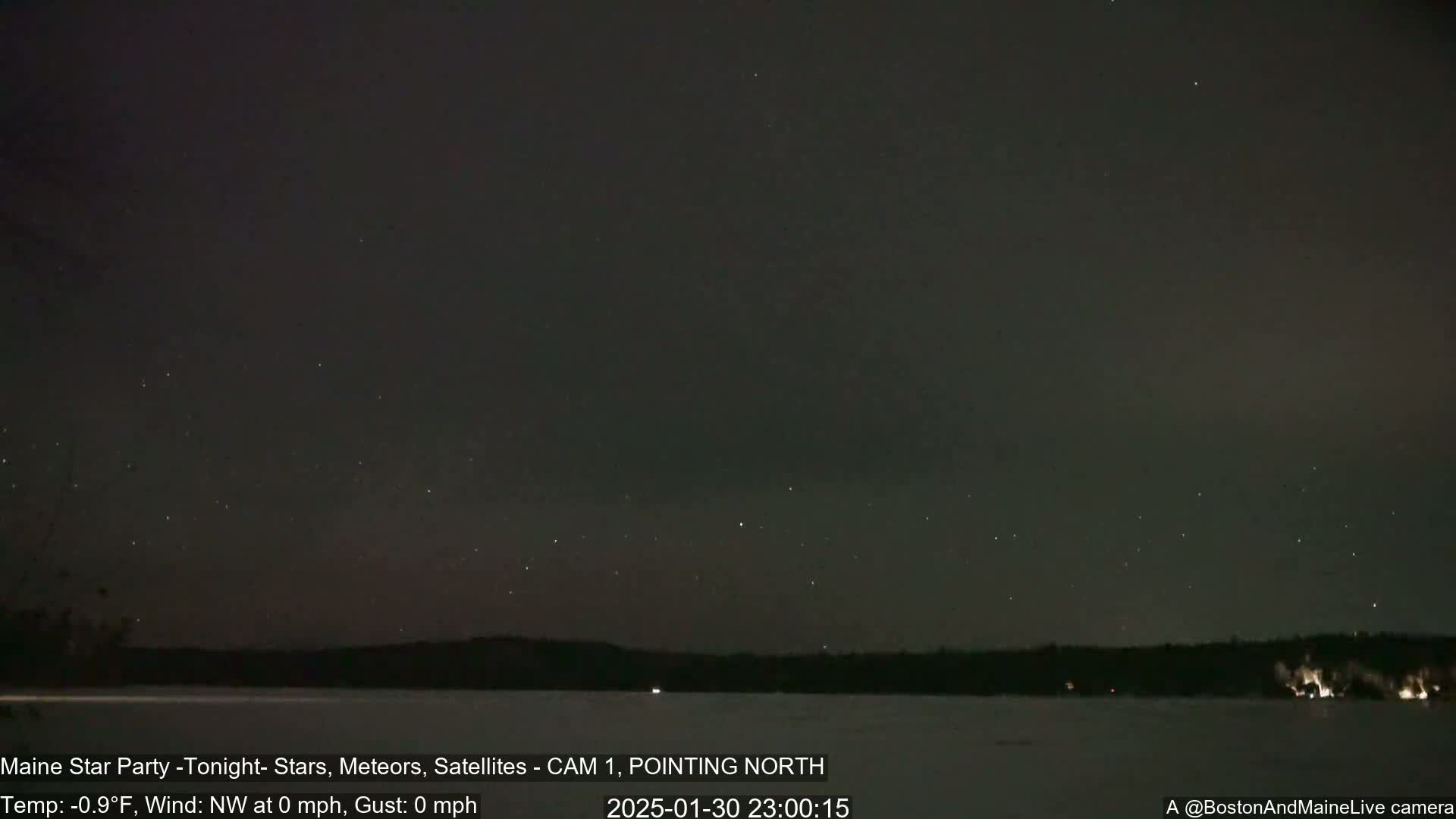 A nighttime view shows numerous stars and a few meteors in a dark sky above a snow-covered lake and dark trees.