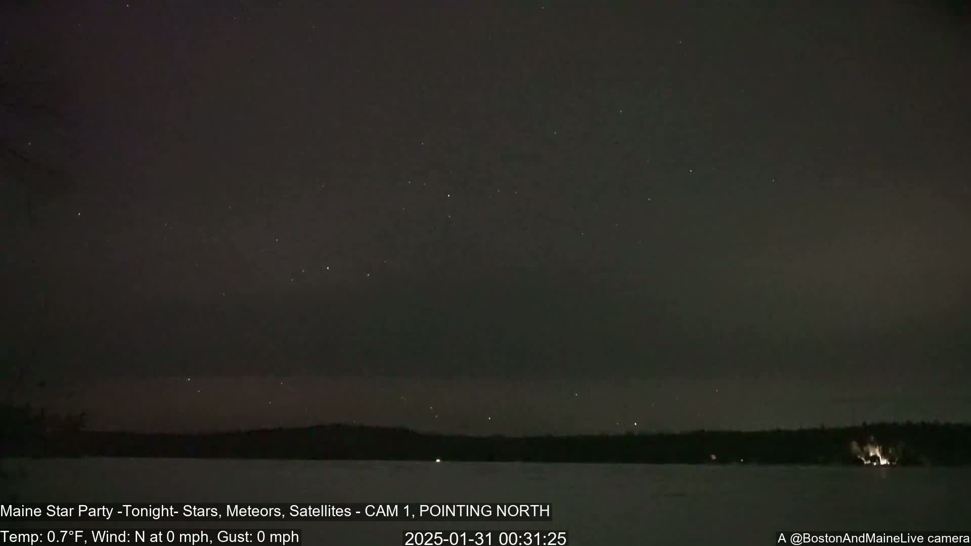A nighttime, still image shows a dark sky filled with stars over a calm, frozen body of water and a dark treeline.